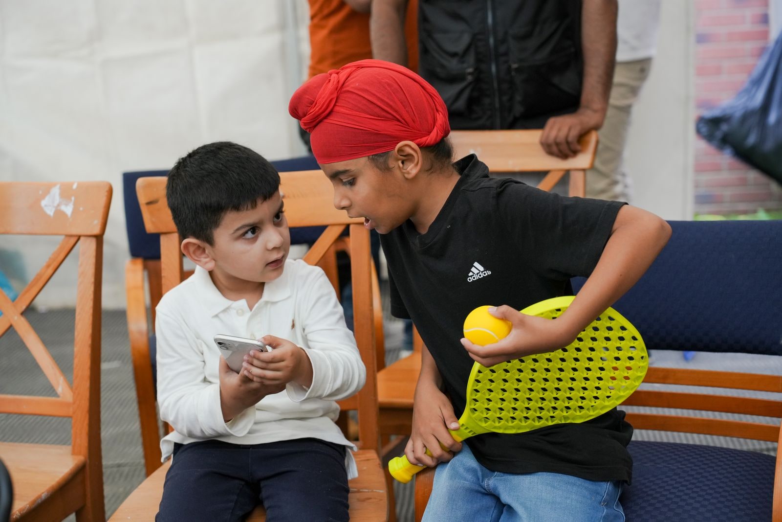 Two boys sitting together at the wedding, one in a red patka holding a bat