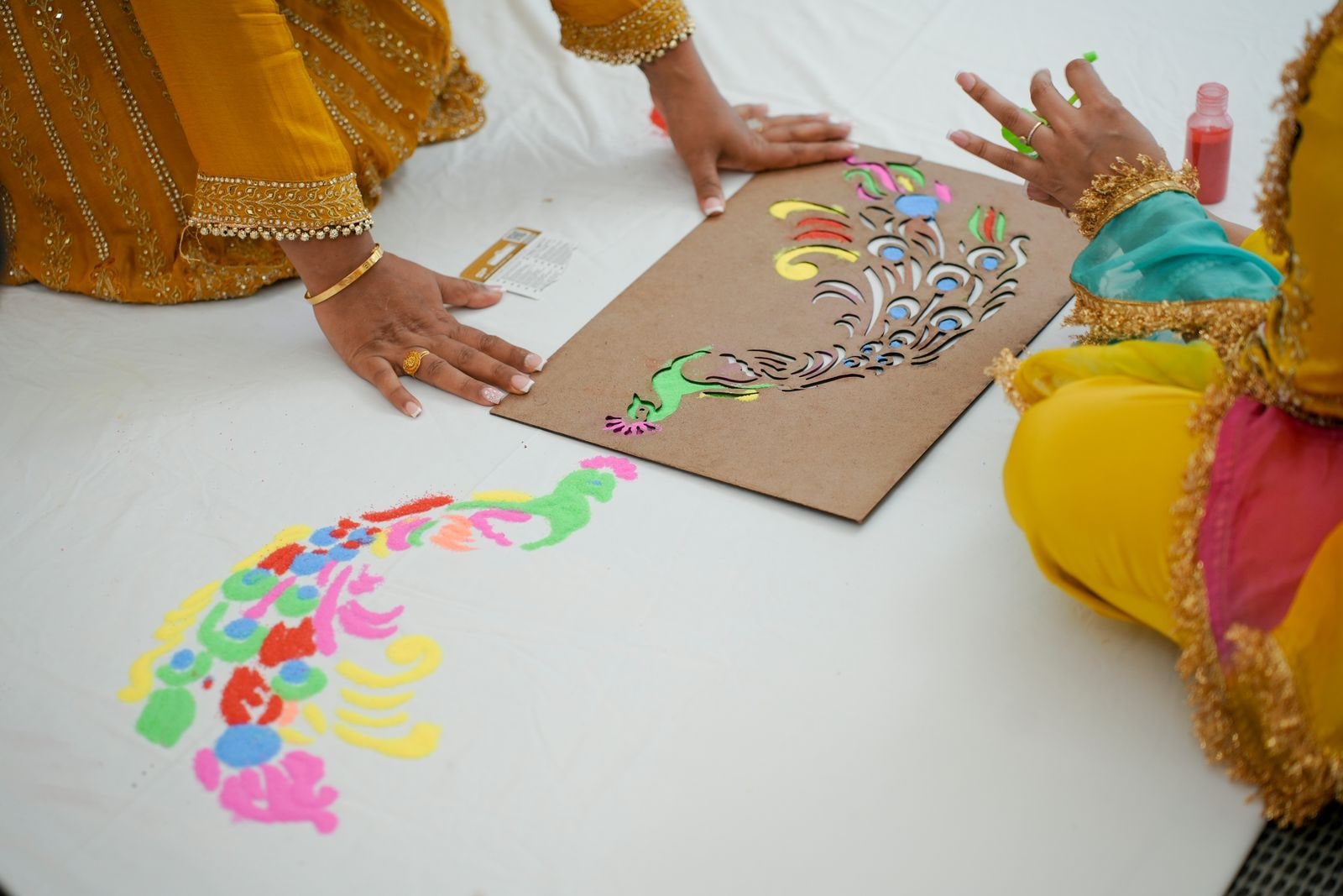 Hands filling in a stencilled rangoli in bright powder on a white sheet