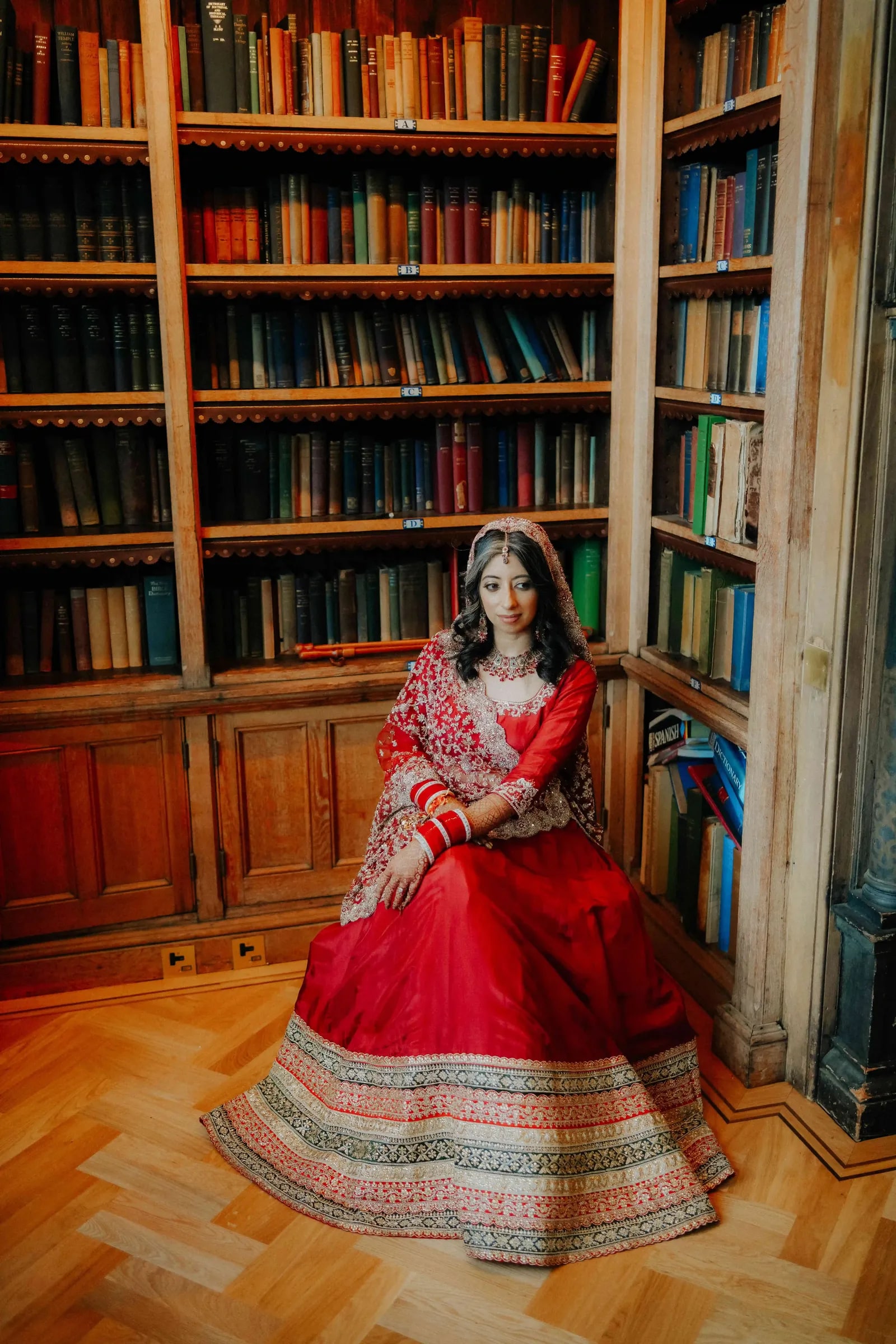Bride seated in a red lehenga against the bookcases of the college library