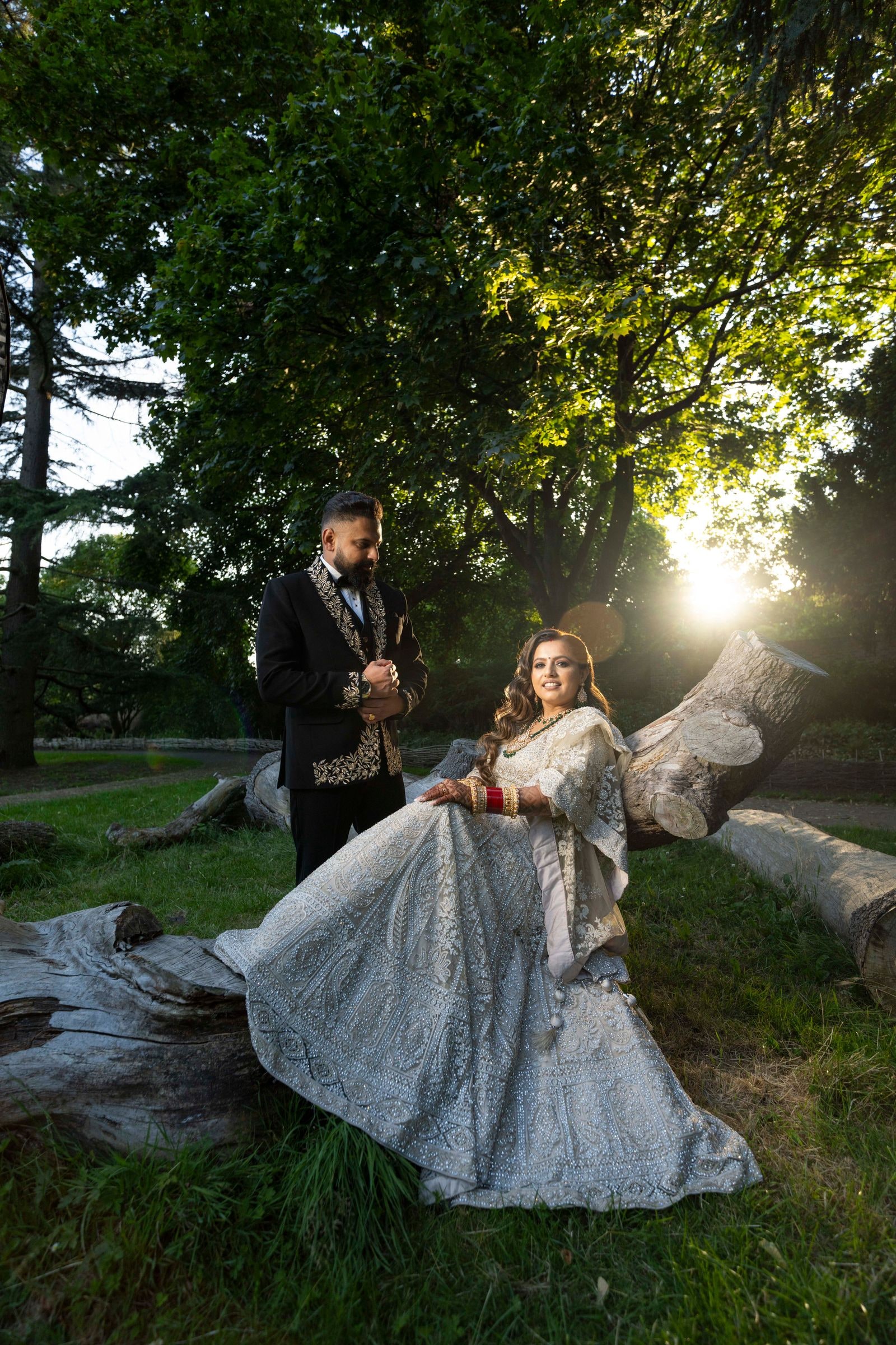 Couple under a tree at sunset, her pale lehenga spread across a fallen trunk