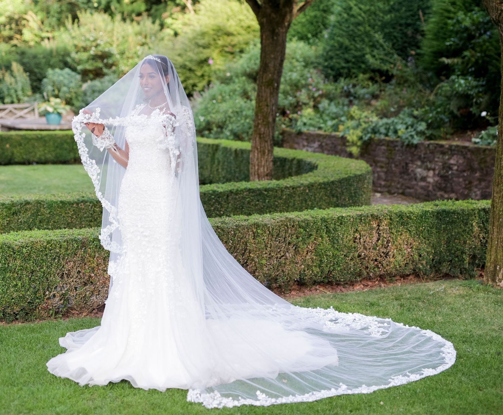 Bride alone on the lawn between clipped hedges, her cathedral veil spread out