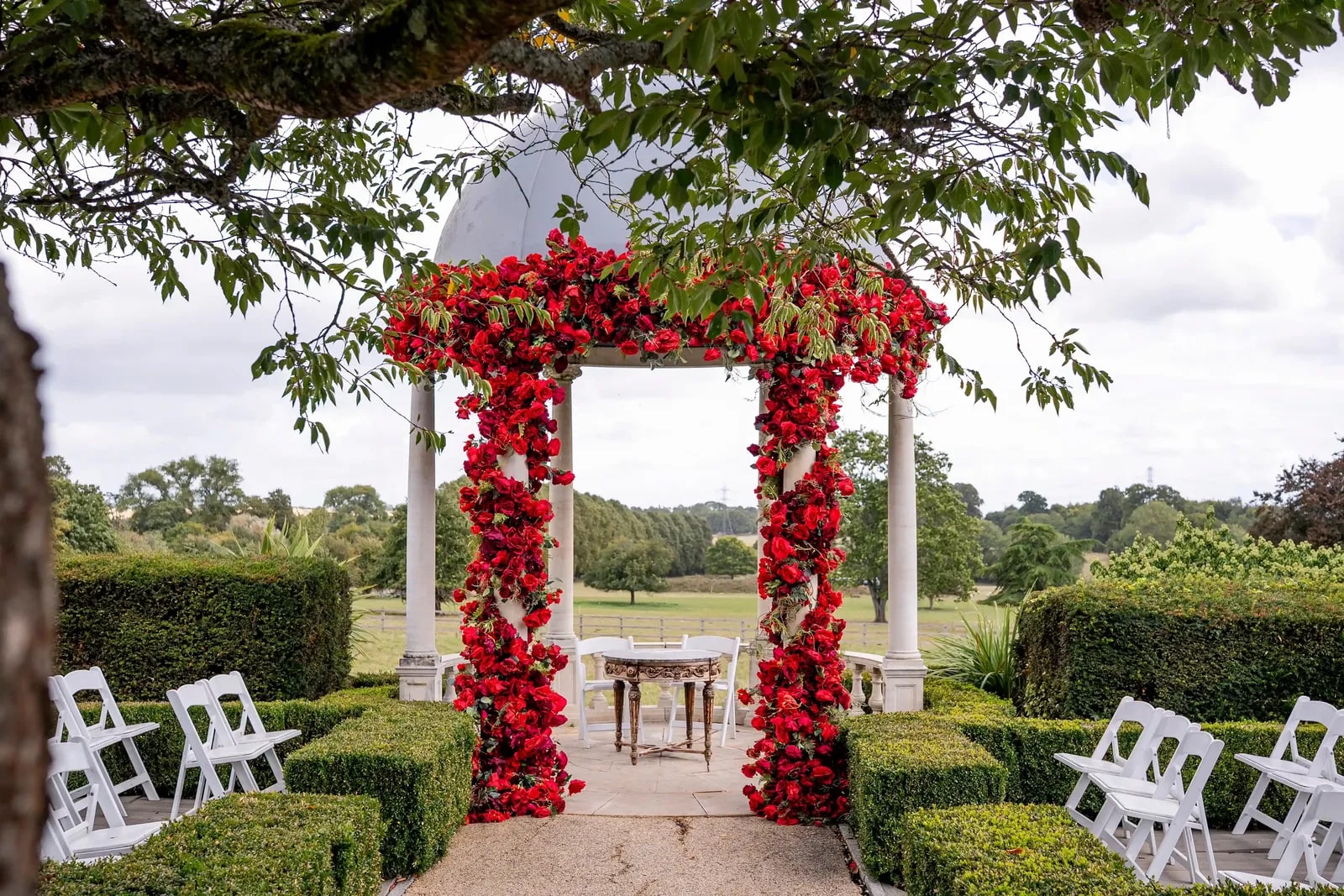 Ceremony arch covered in red roses, set out with white chairs looking over the fields