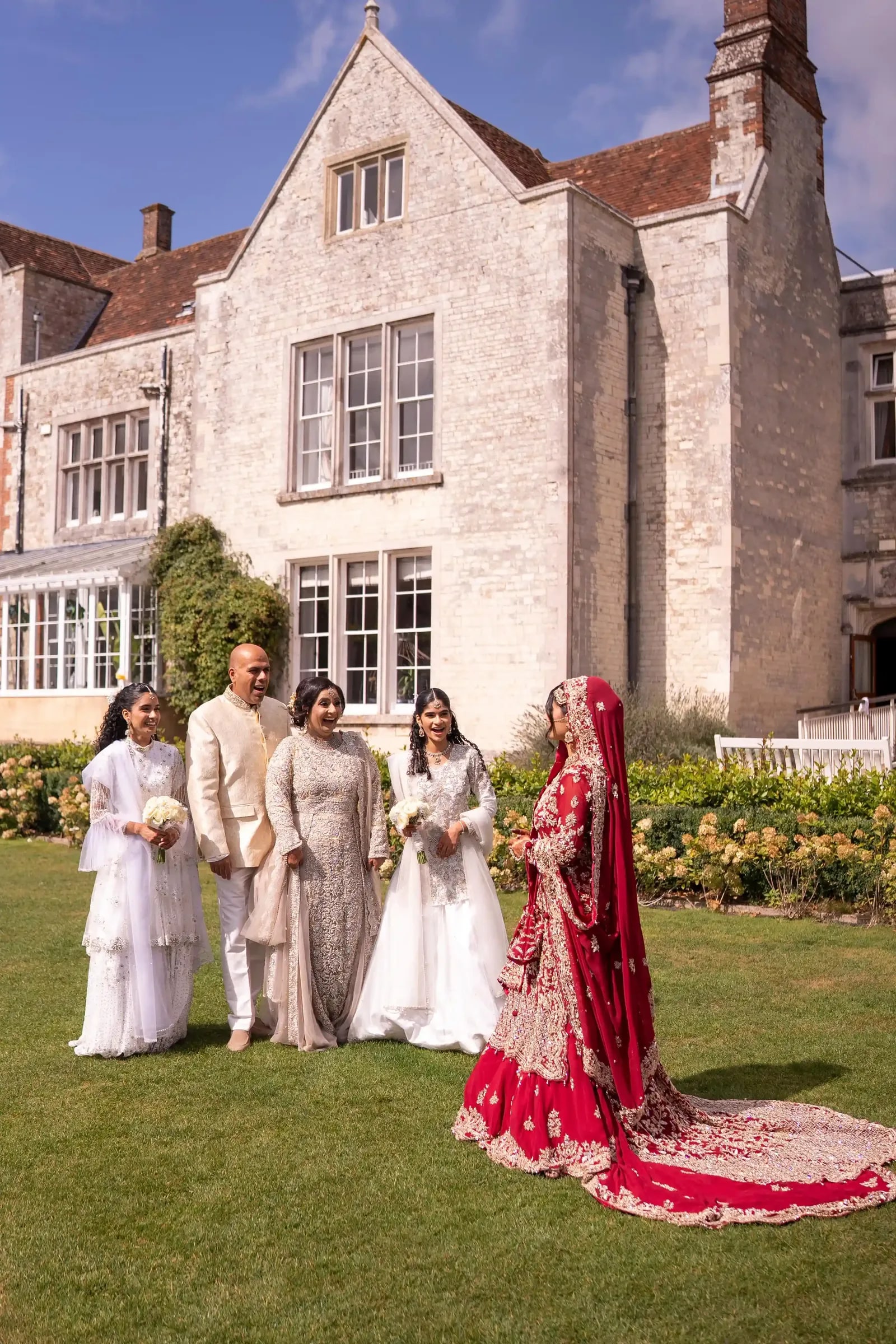 Bride in crimson standing apart from her family in ivory on the lawn
