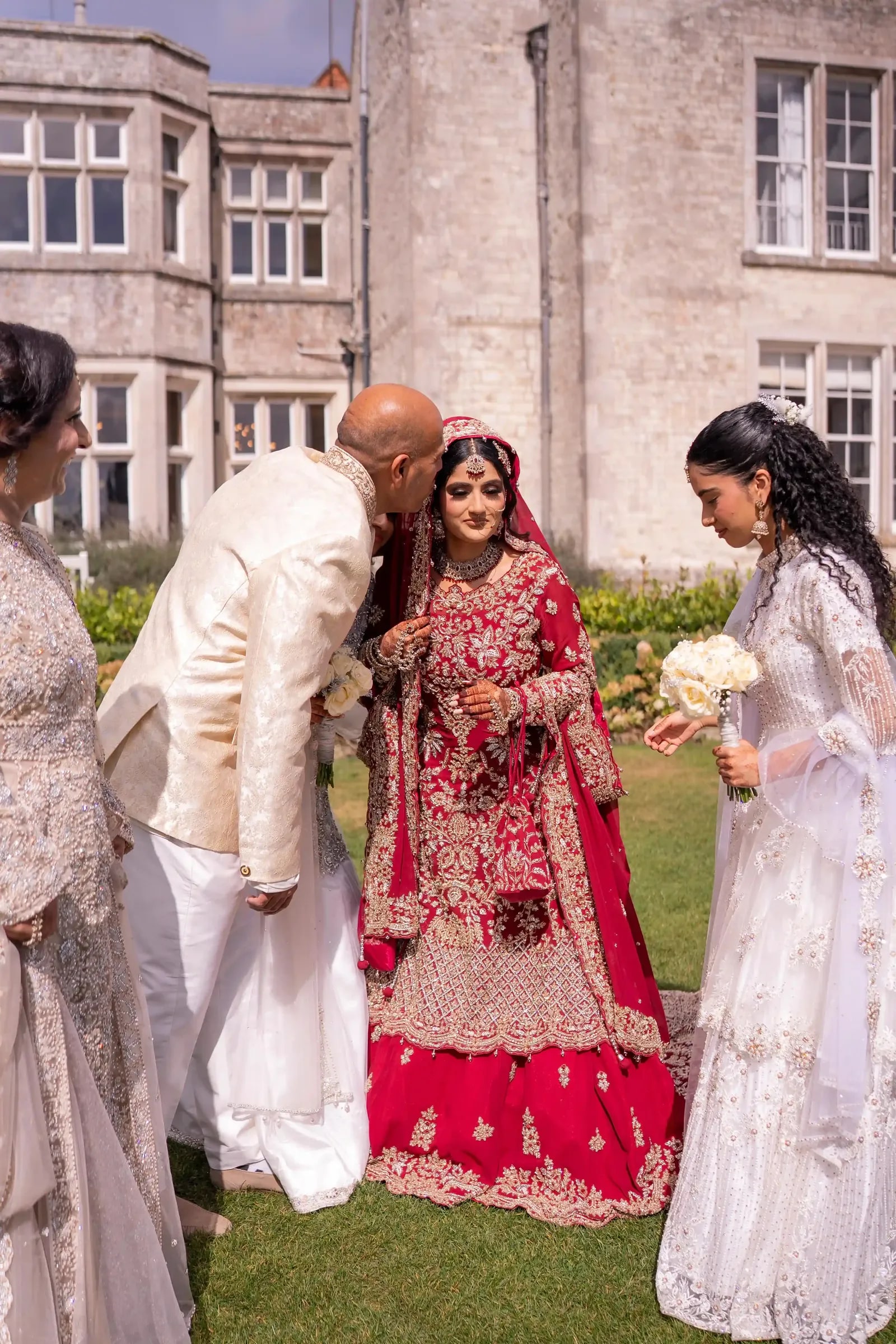 The bride's father kissing her forehead on the lawn, her sisters beside them