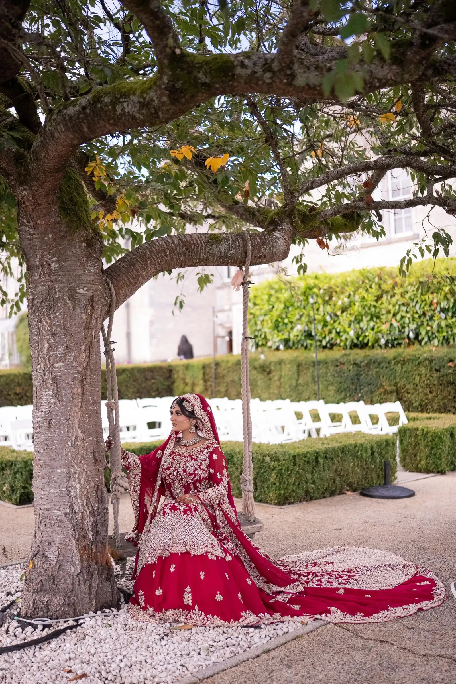 Bride seated alone under a spreading tree, her crimson train laid across the gravel