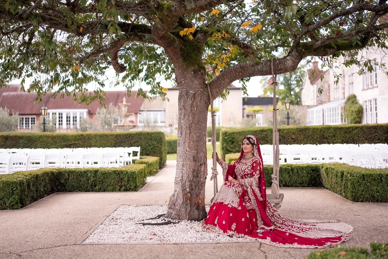 Bride on a swing beneath a tree in the garden, her crimson train across the gravel