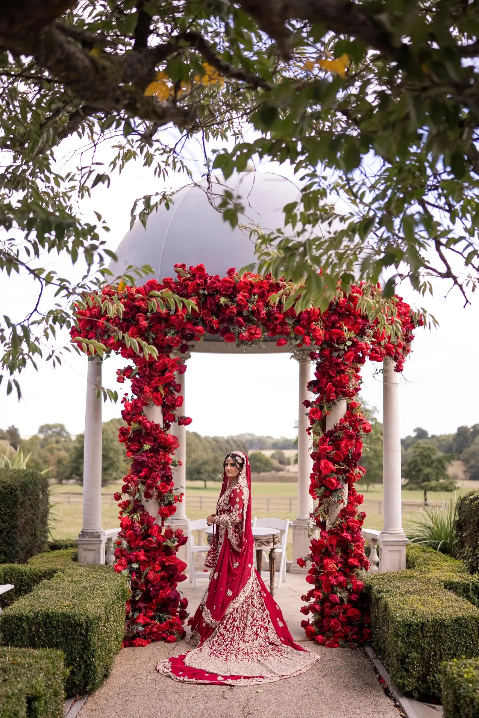 Bride standing under the red rose arch, the fields beyond stretching away