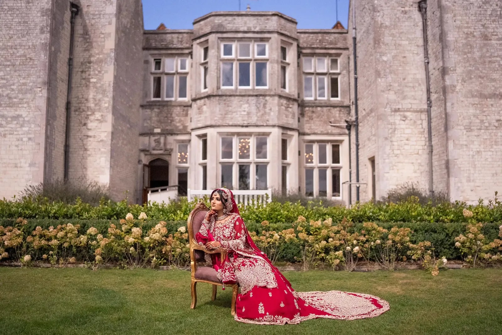 Bride seated on a gilt chair on the lawn in front of the house, her train spread out