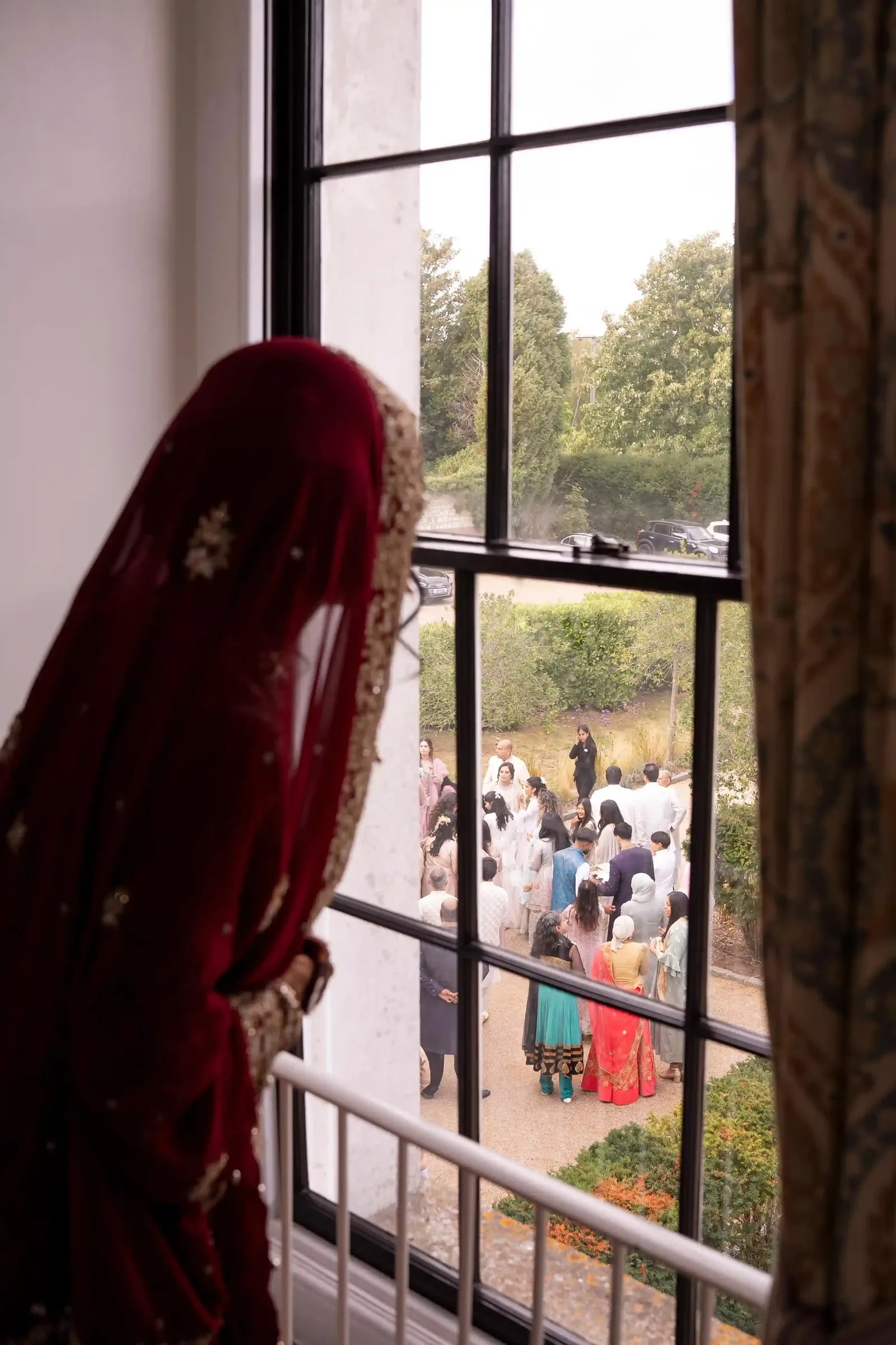 Bride watching the guests gather below from a tall window, seen from behind