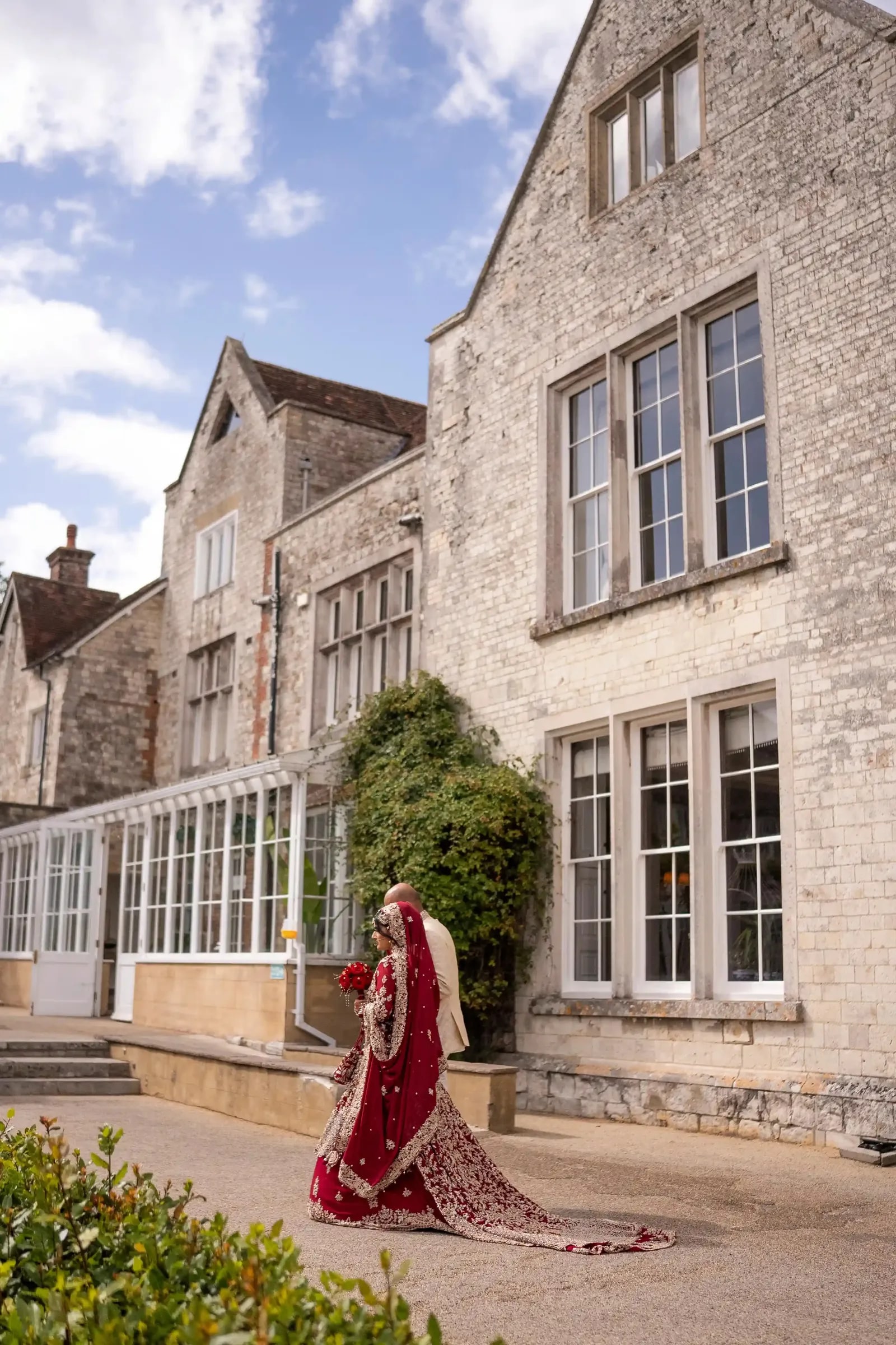 Bride in crimson alone at the corner of the house, her train trailing on the paving