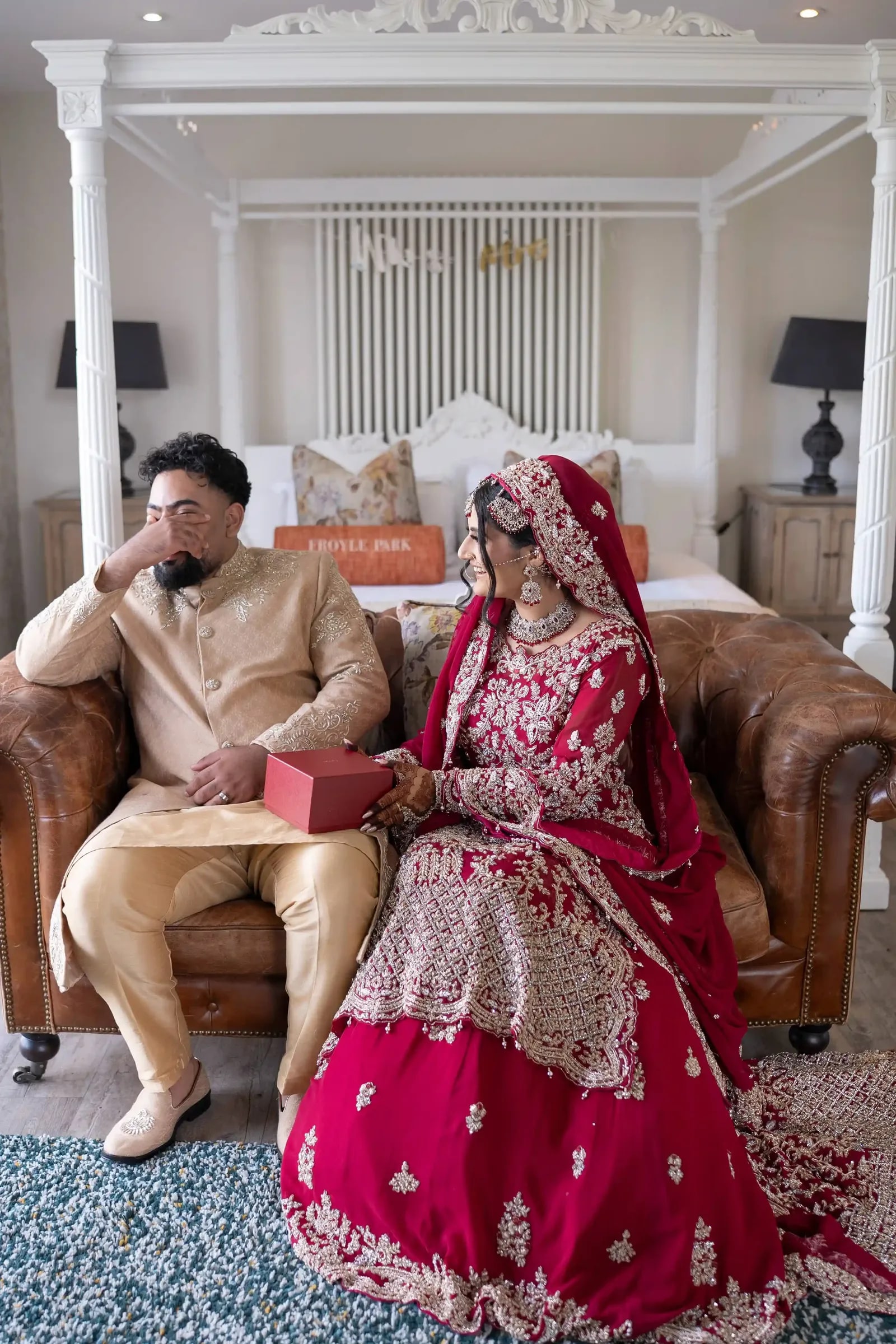 Bride and groom on a leather sofa in the suite, he holding a red gift box