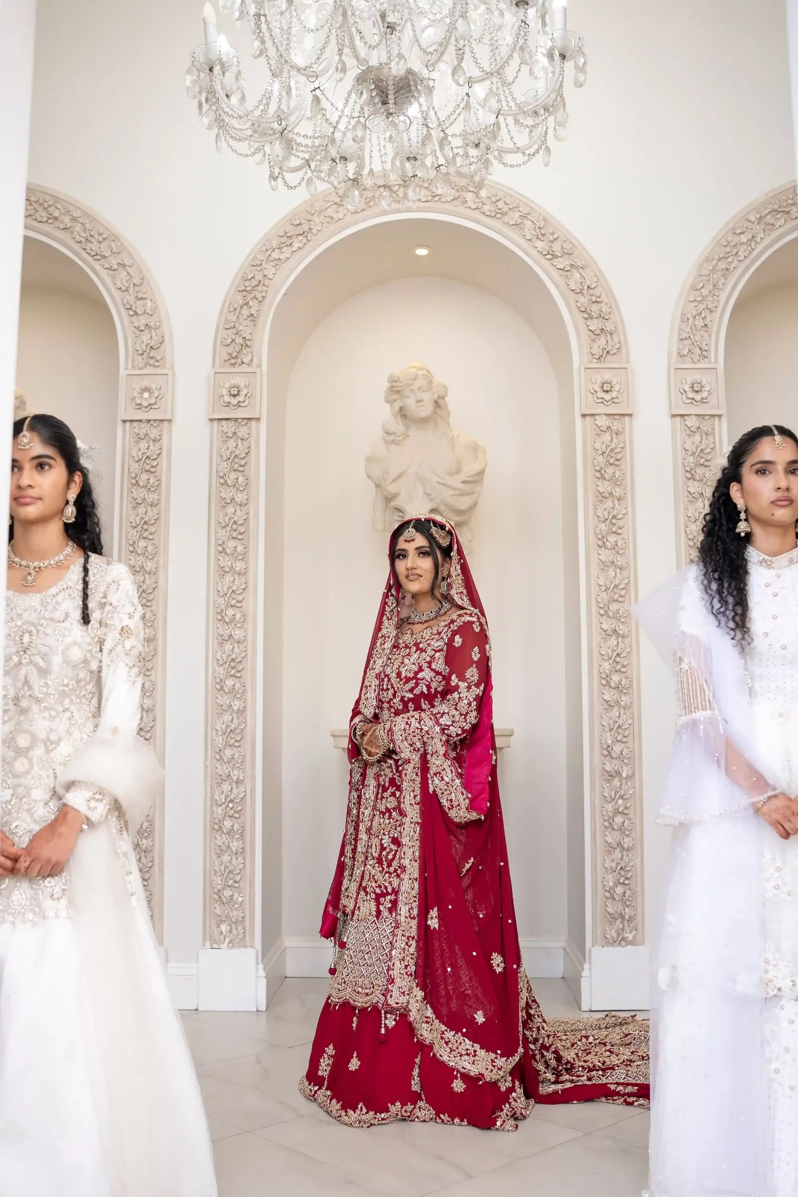 Bride in crimson between two women in ivory, framed by white plaster arches