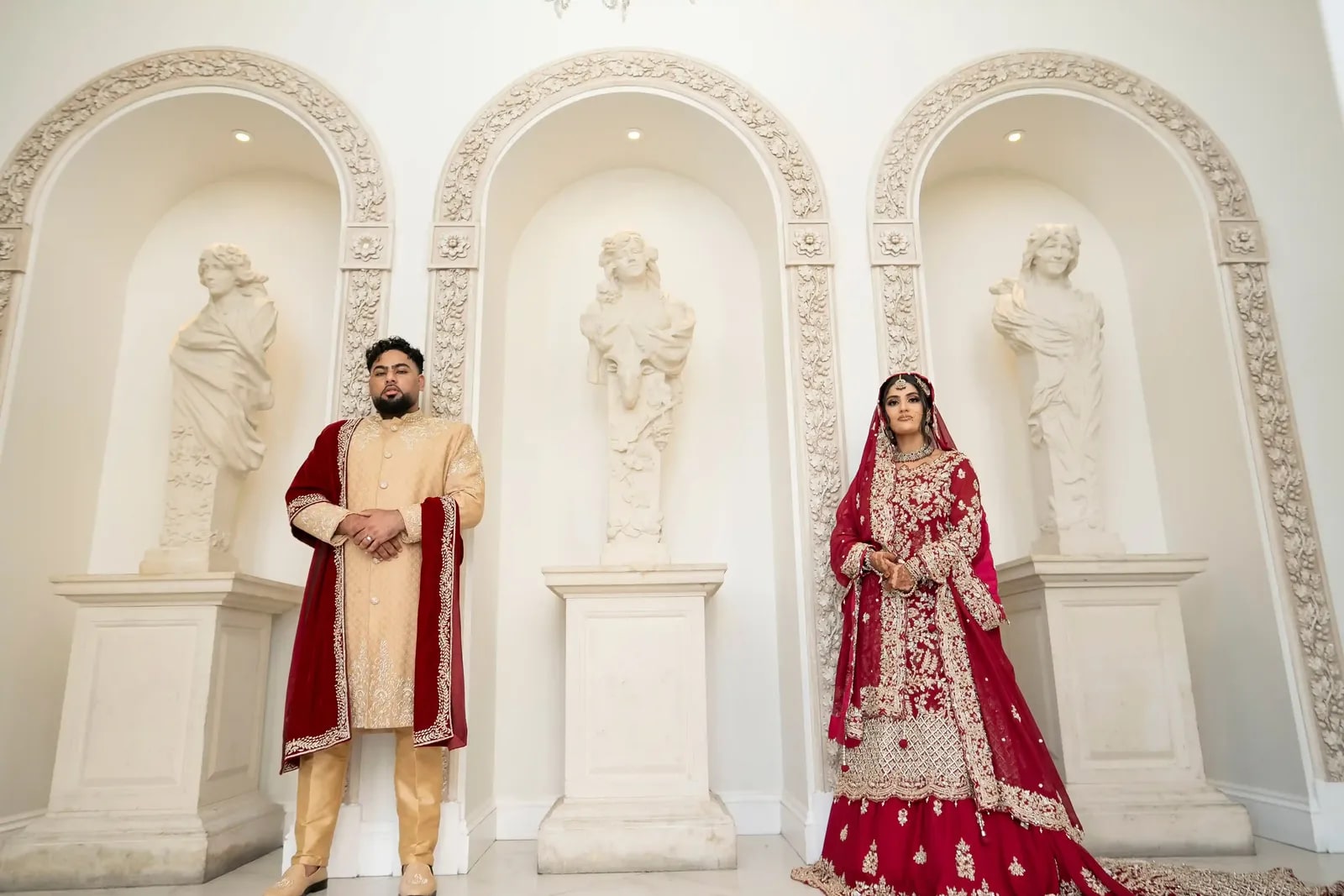 Bride and groom standing apart in front of white statuary in alcoves