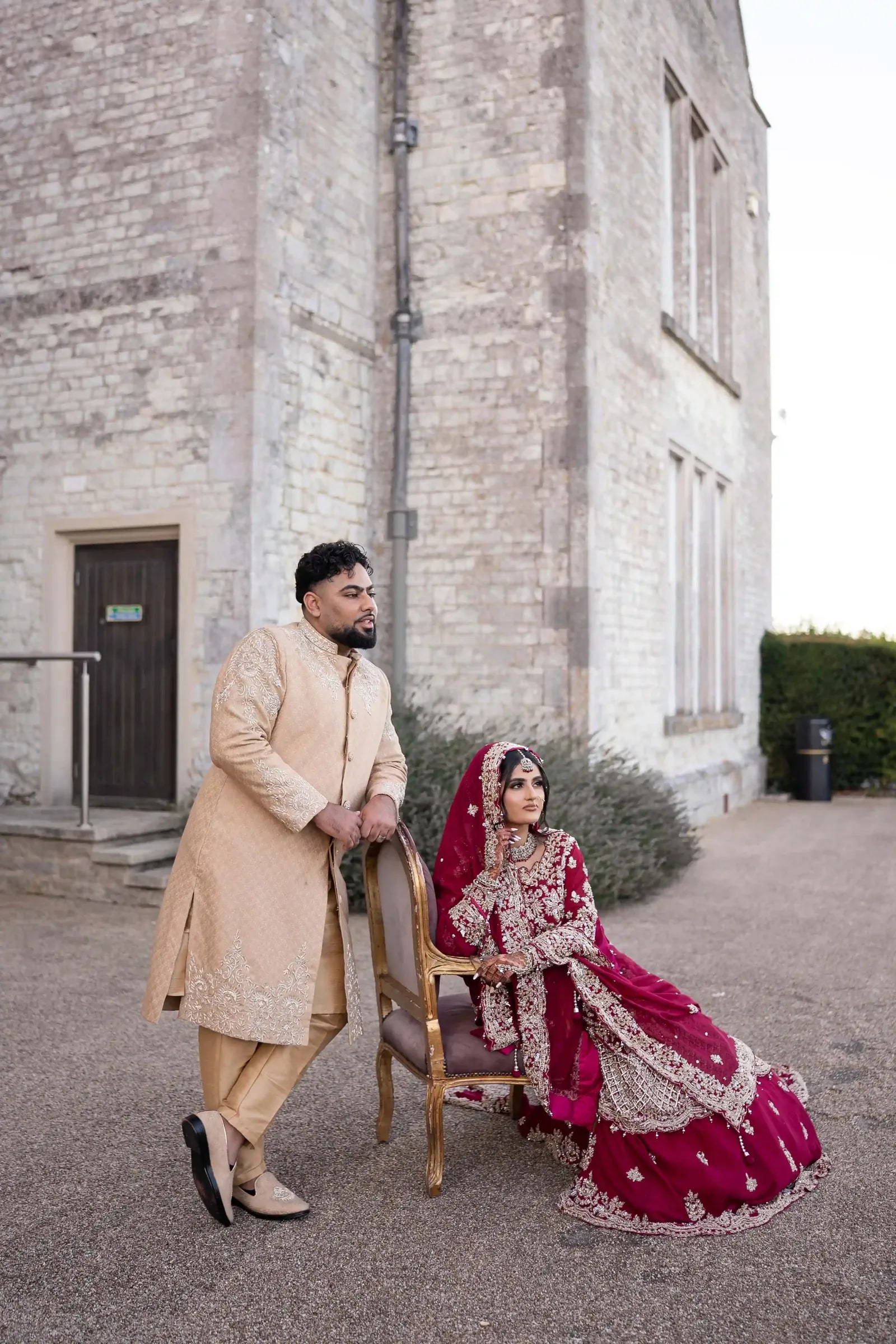 Groom leaning on a chair where his bride sits against the stone wall of the house