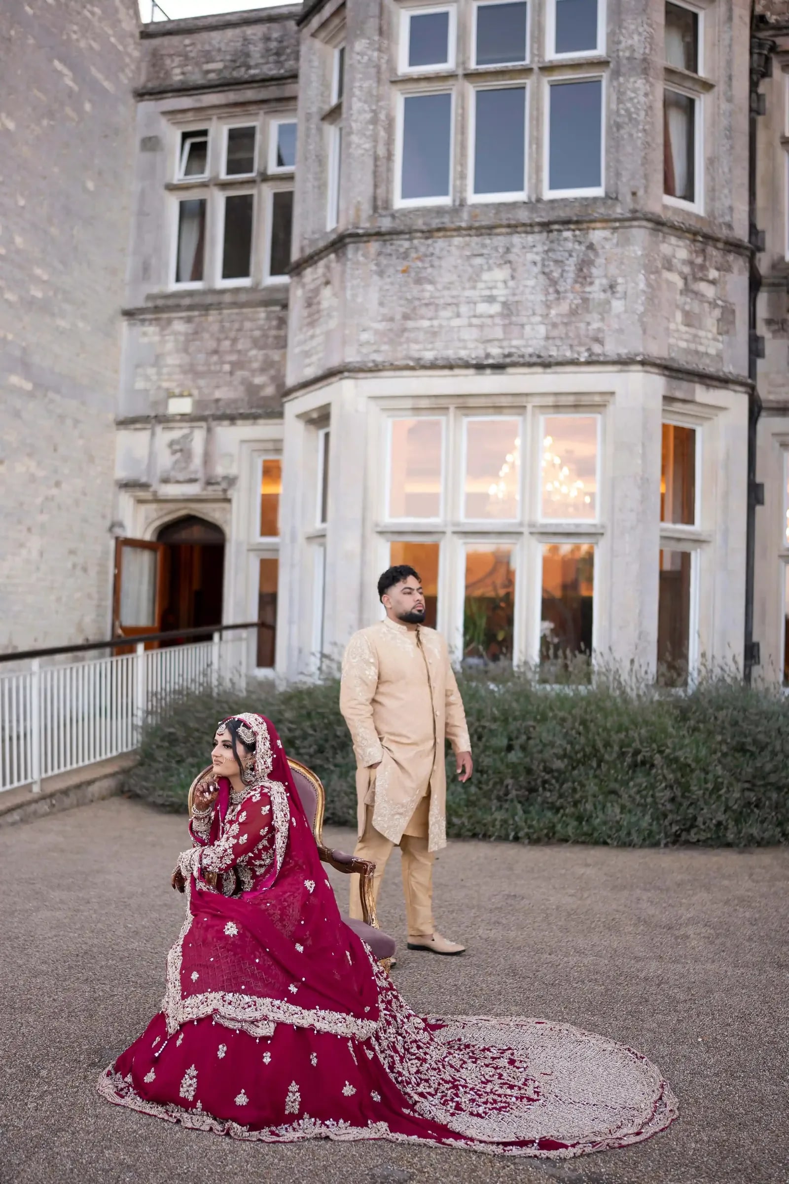Bride seated on the gravel in crimson, her groom standing behind her at dusk