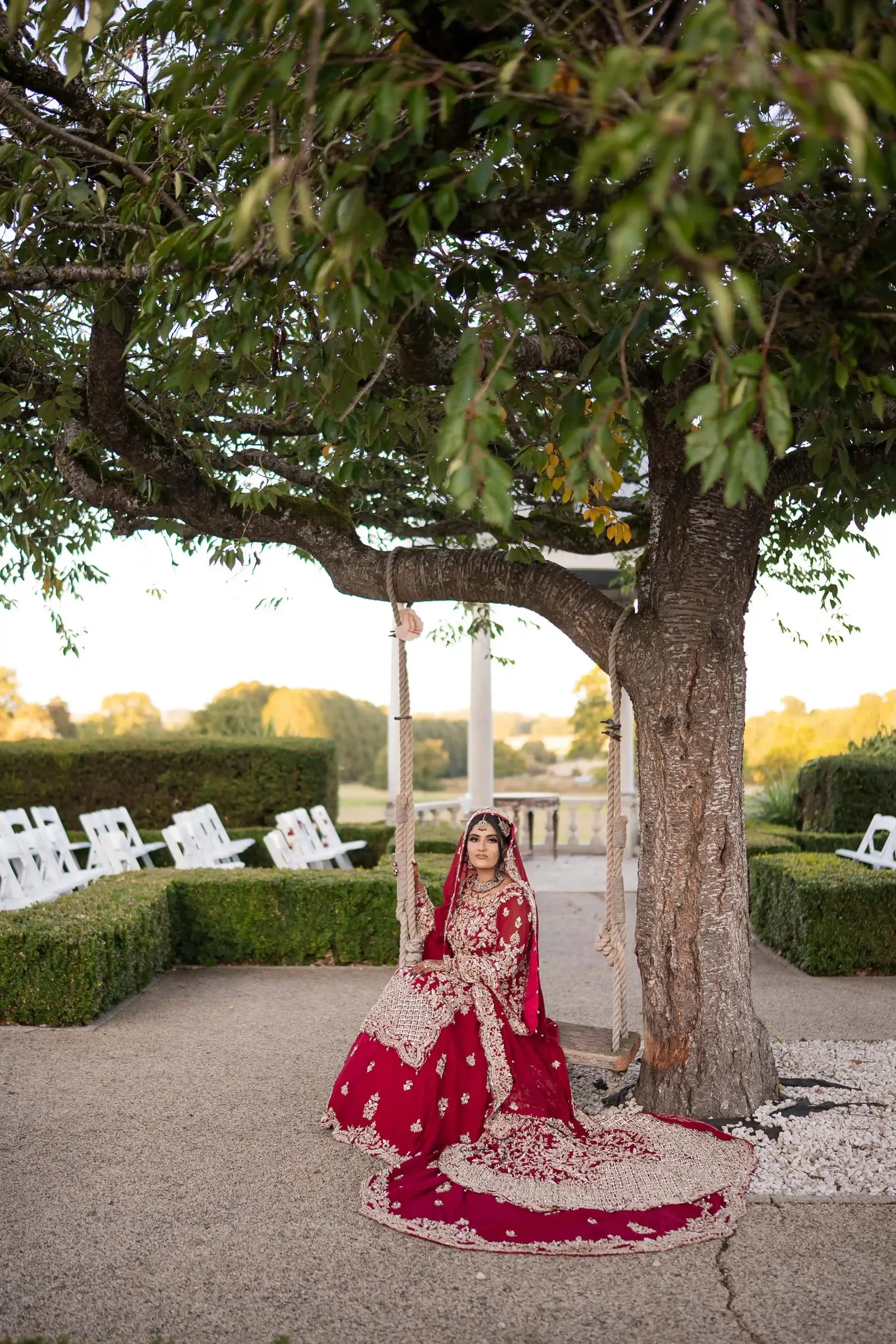 Bride on the swing beneath the tree at golden hour, her train laid across the gravel