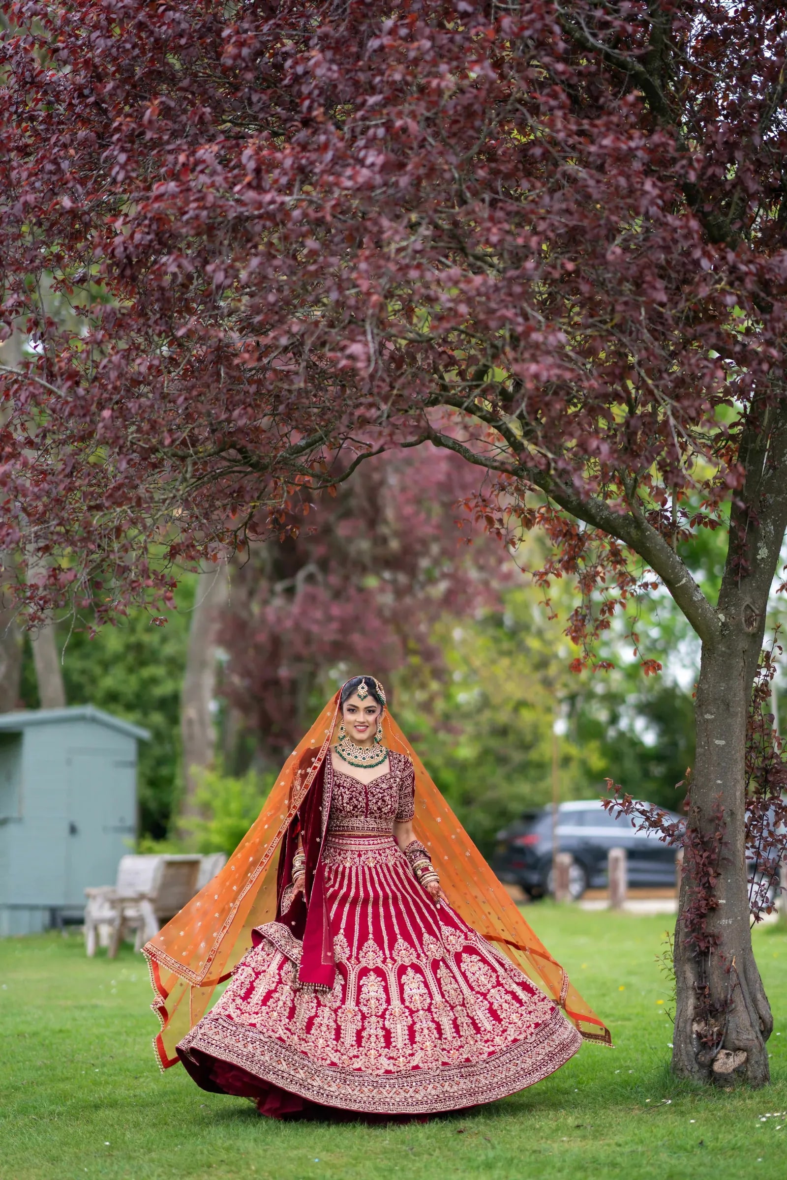 Bride turning beneath a copper beech, her red lehenga spread wide