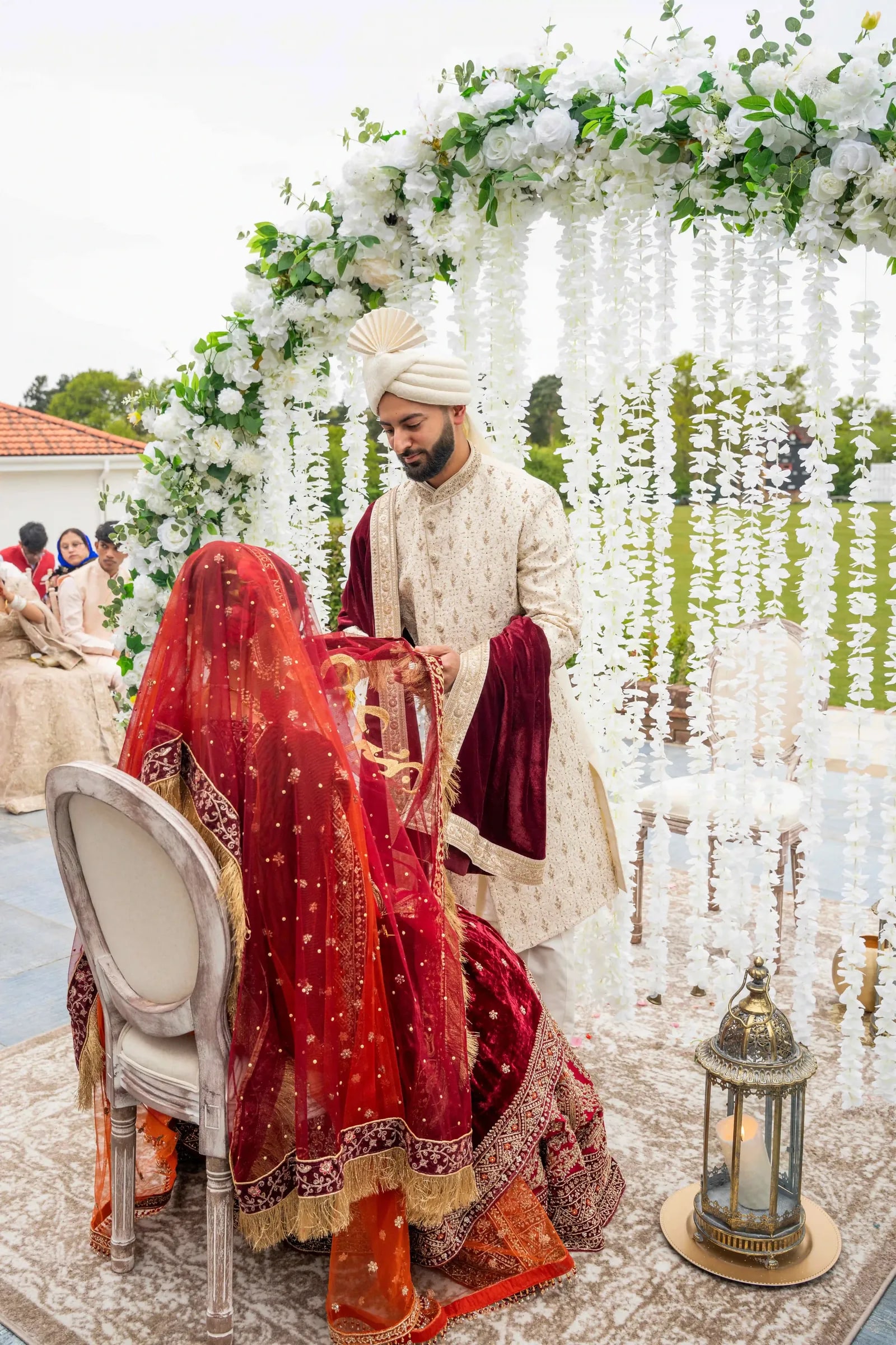 Groom lifting his bride's veil where she sits beneath the hanging white flowers