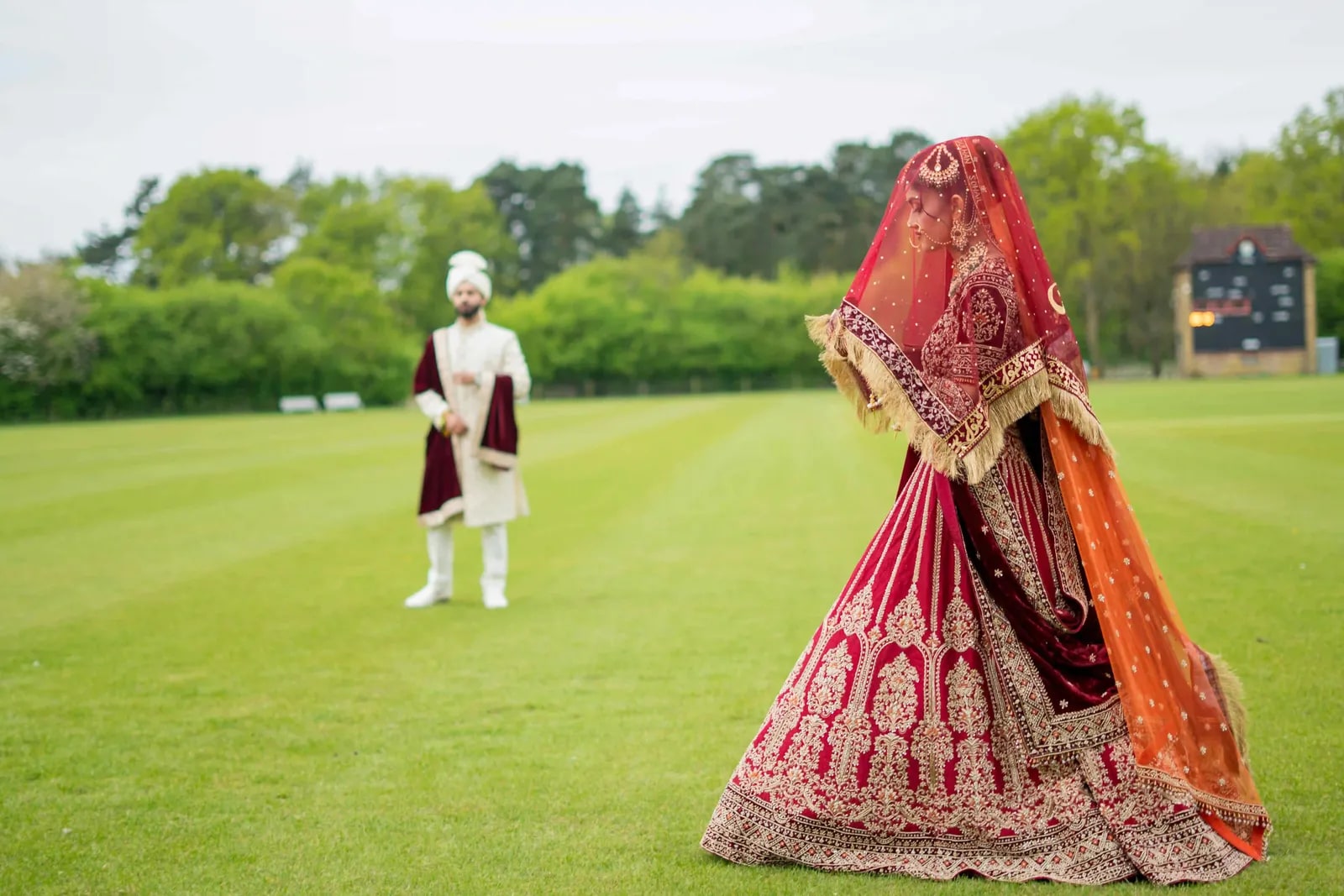 Bride turning on the cricket outfield, her groom standing far behind her
