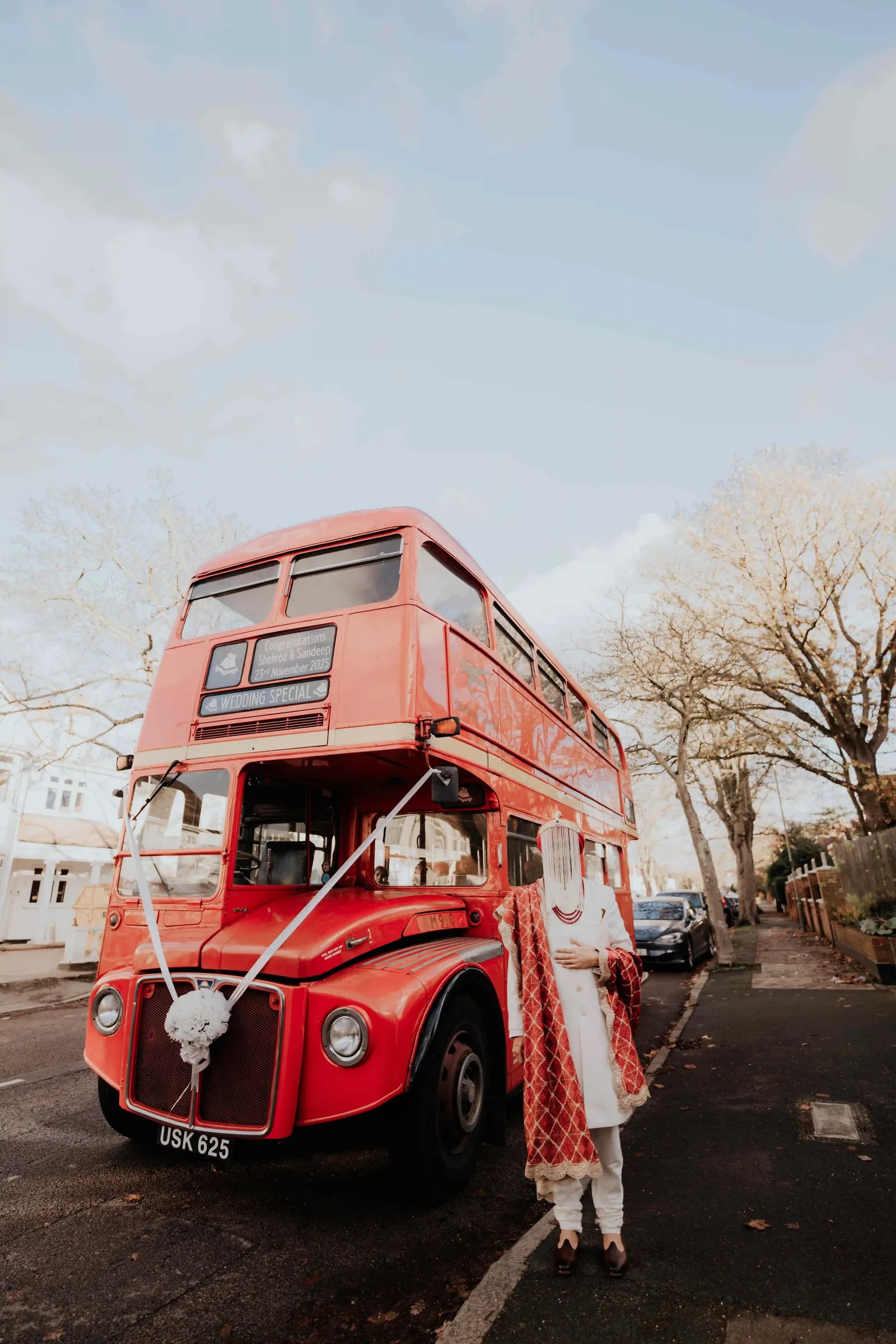 A red Routemaster bus dressed with ribbons, the groom standing beside it