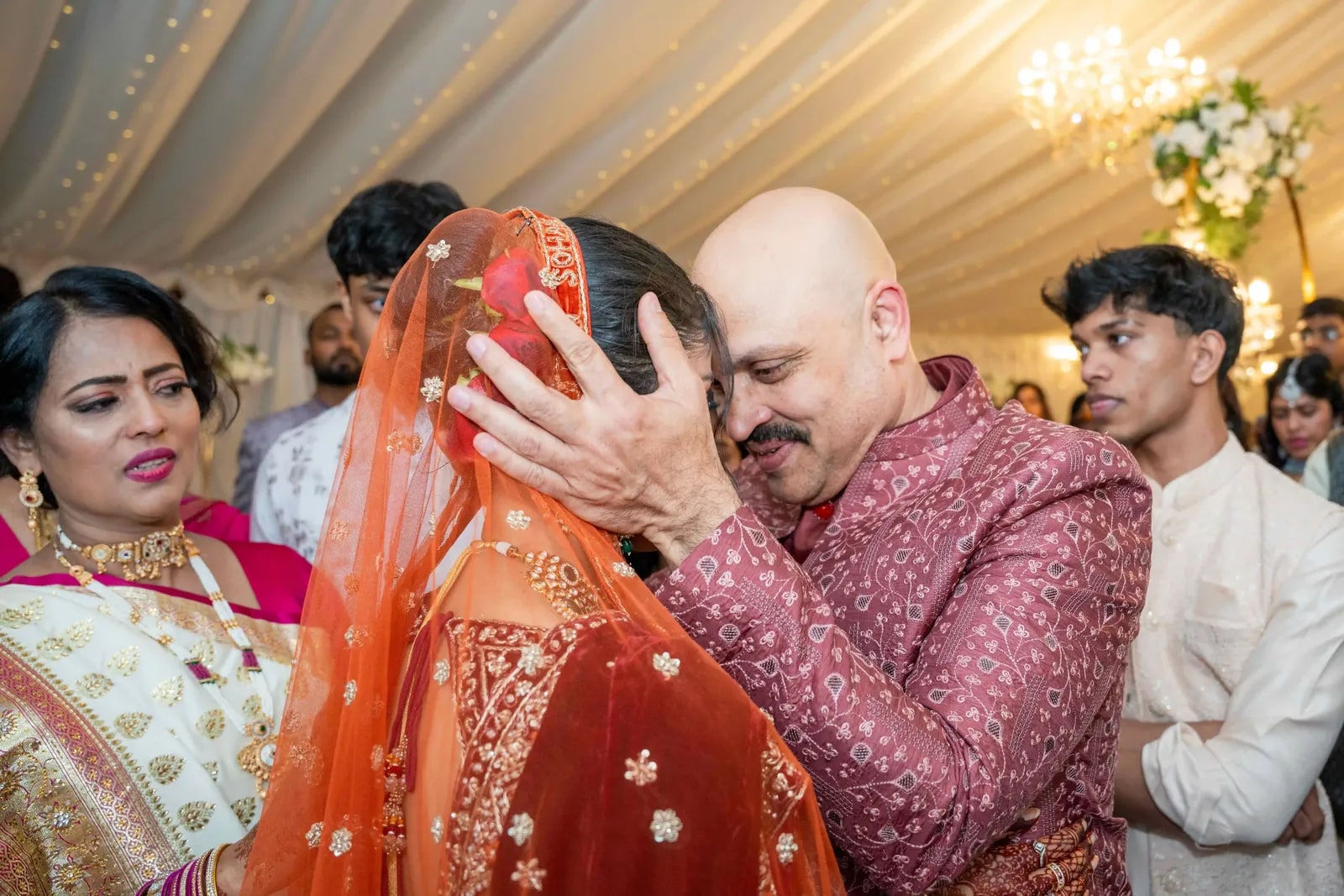 A relative kissing the bride's forehead through her orange dupatta