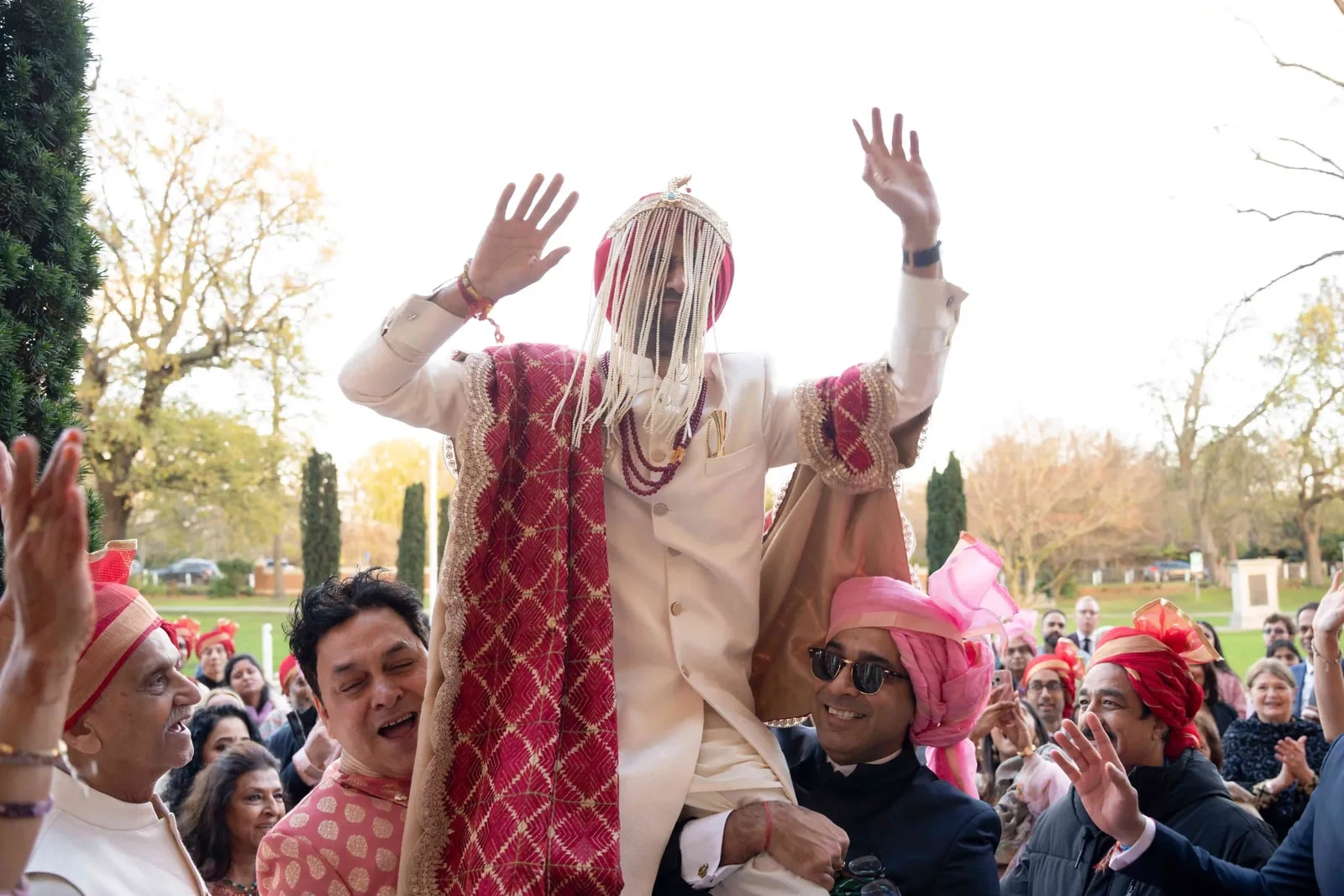 Groom in a sehra with his arms raised, carried through the crowd