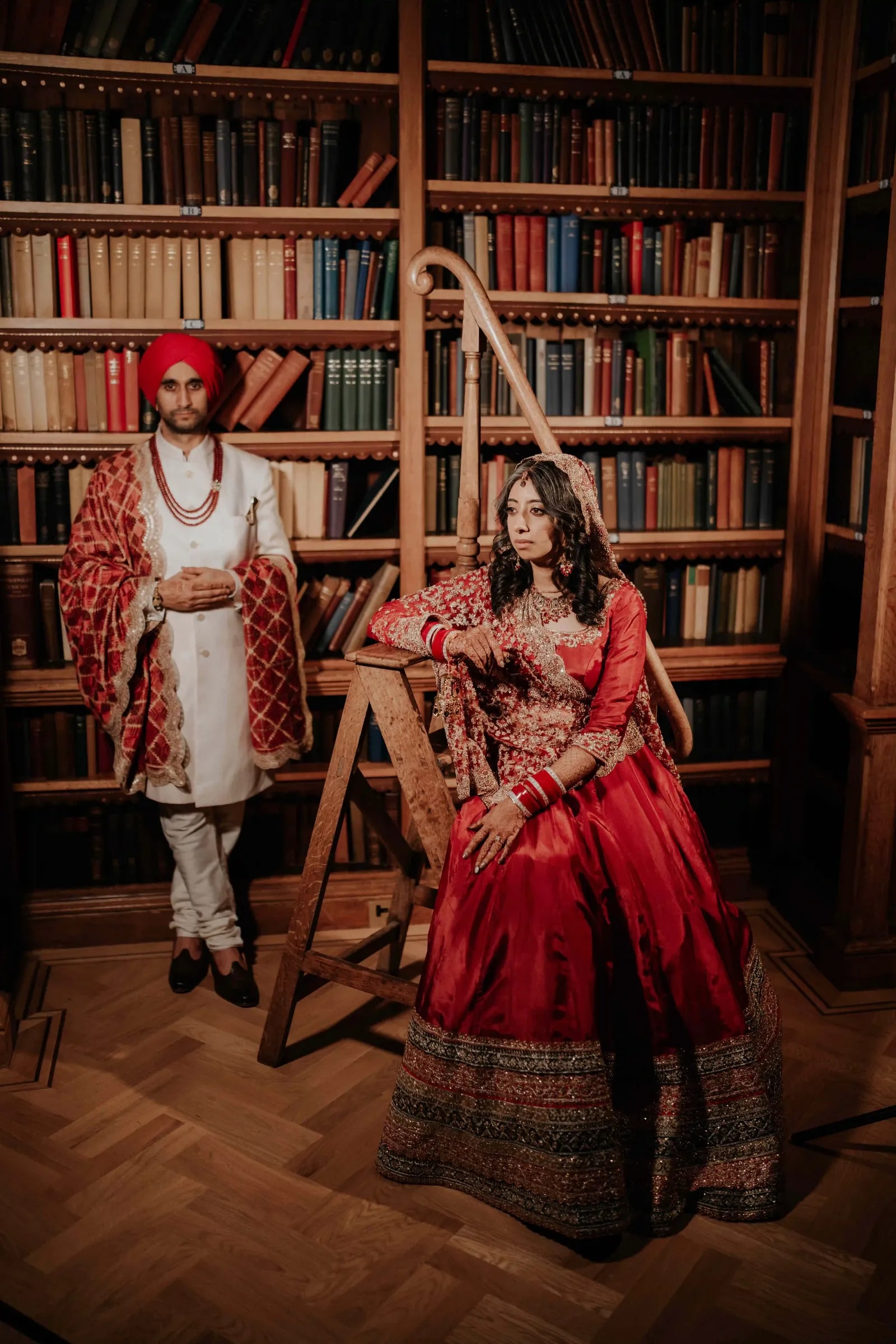 Bride seated by a library ladder in a red lehenga, her groom standing behind