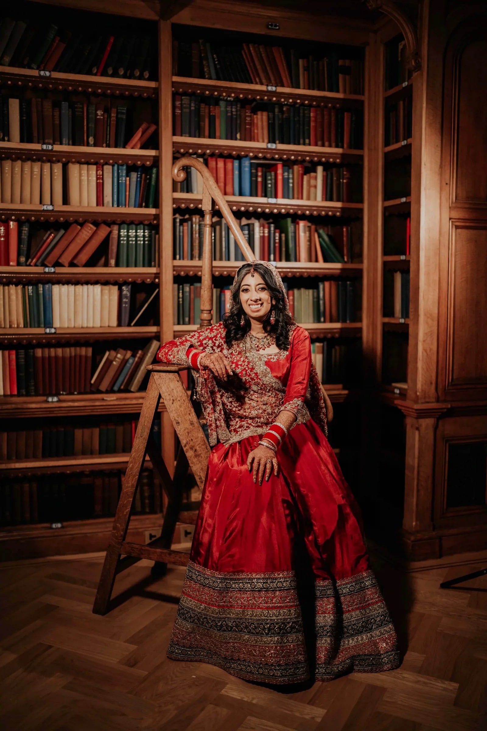 Bride leaning on a library ladder among the bookshelves in a red lehenga