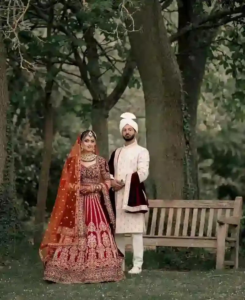 Bride and groom standing by a bench under old trees, she in red, he in ivory and burgundy