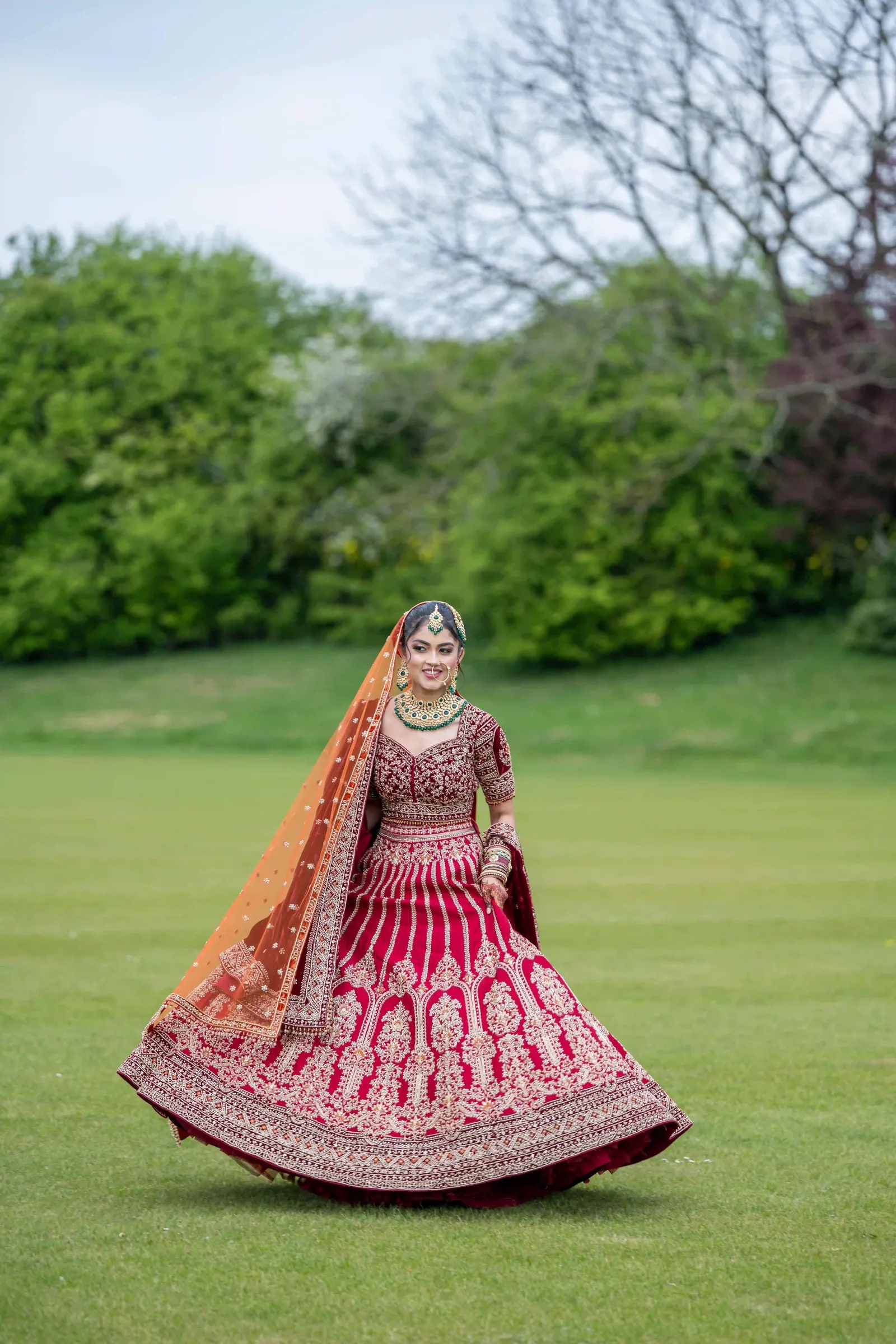 Bride walking alone across the lawn in a deep red lehenga and orange dupatta