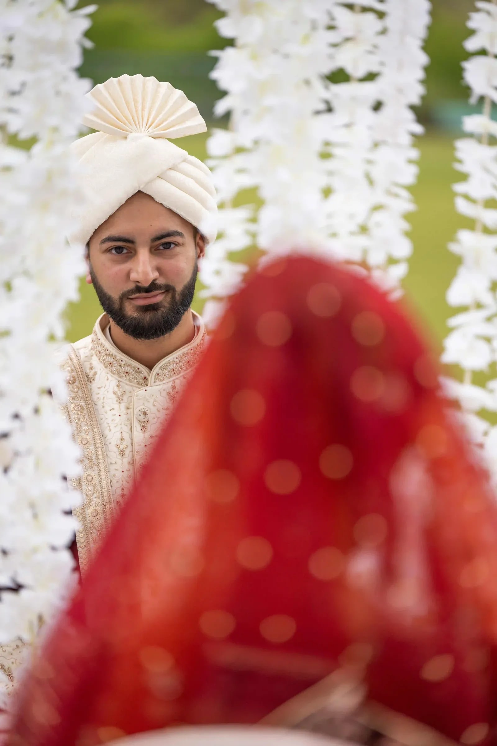 Groom in an ivory turban seen past the blurred red of his bride's dupatta