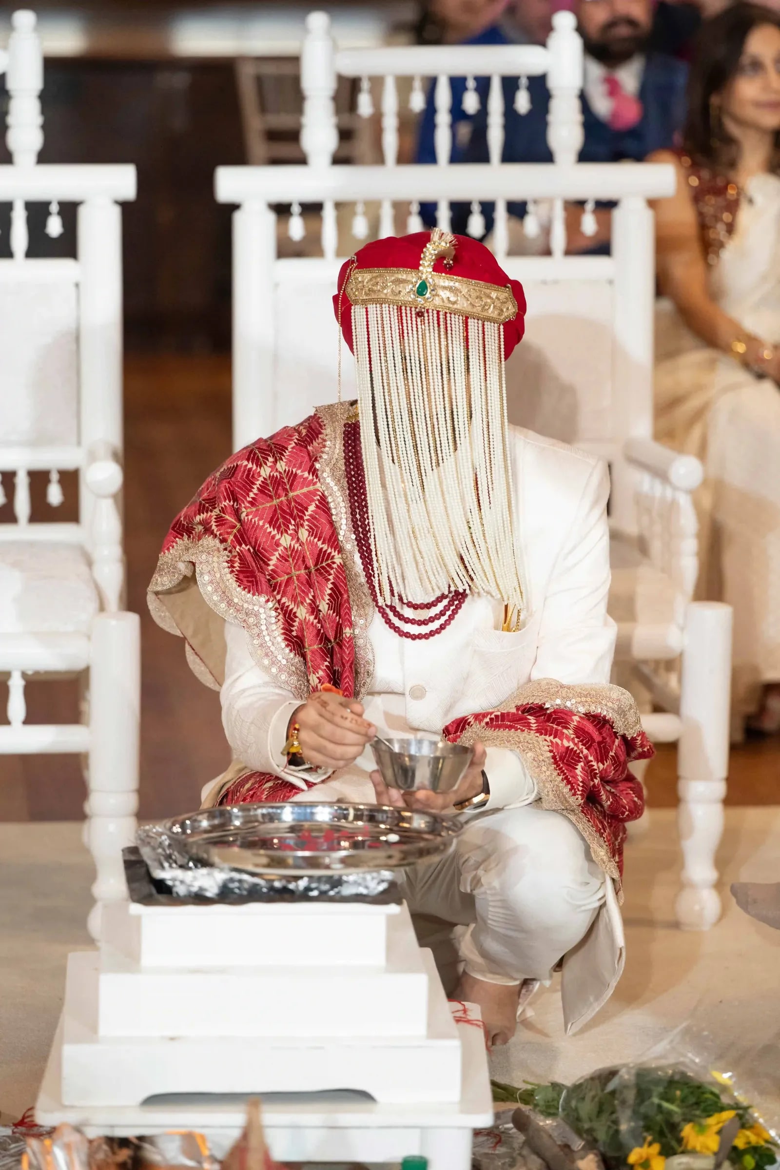 Groom in a red turban and sehra of strung beads seated at the ceremony