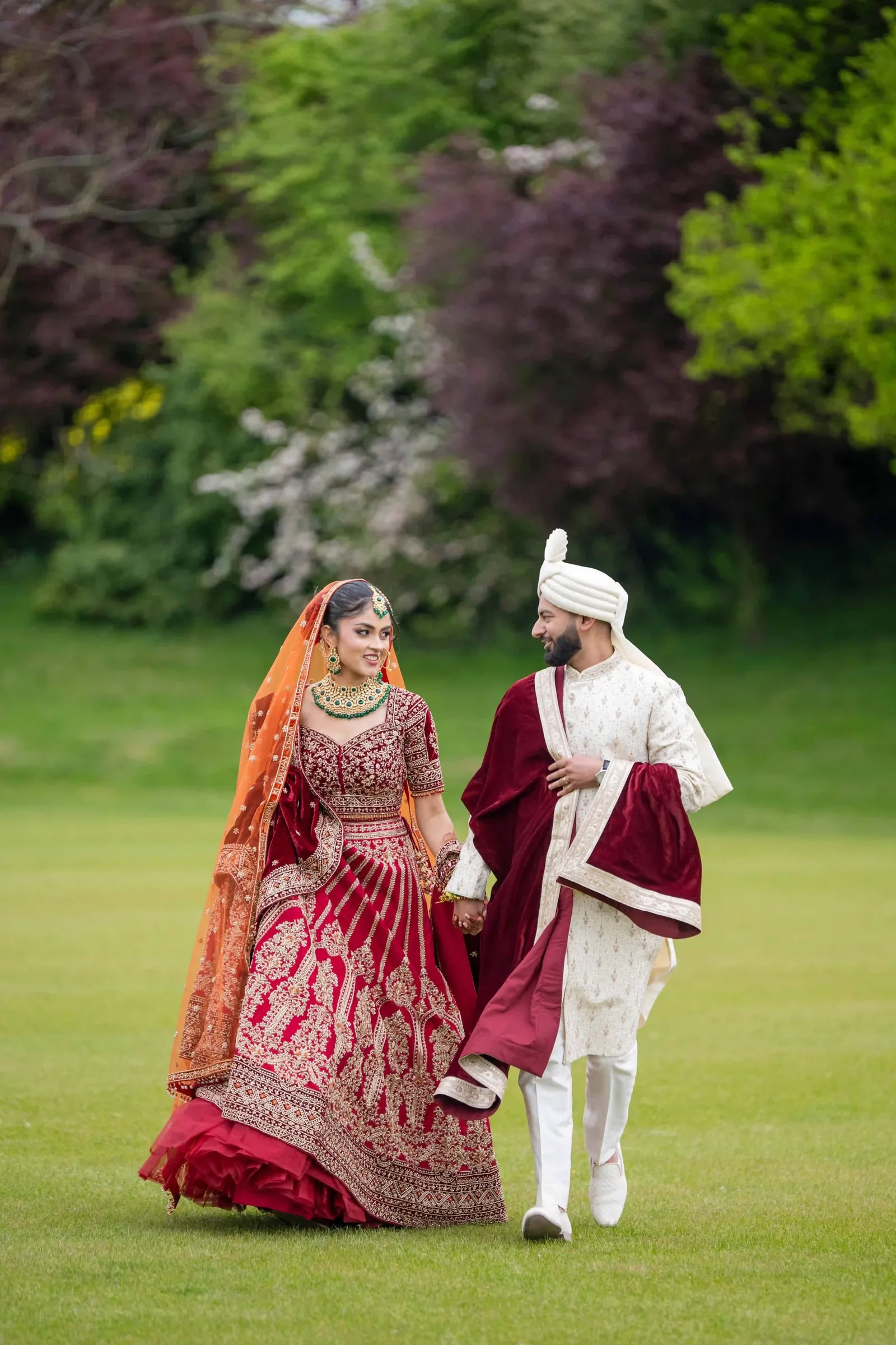Bride and groom walking hand in hand across the lawn, he in ivory and burgundy