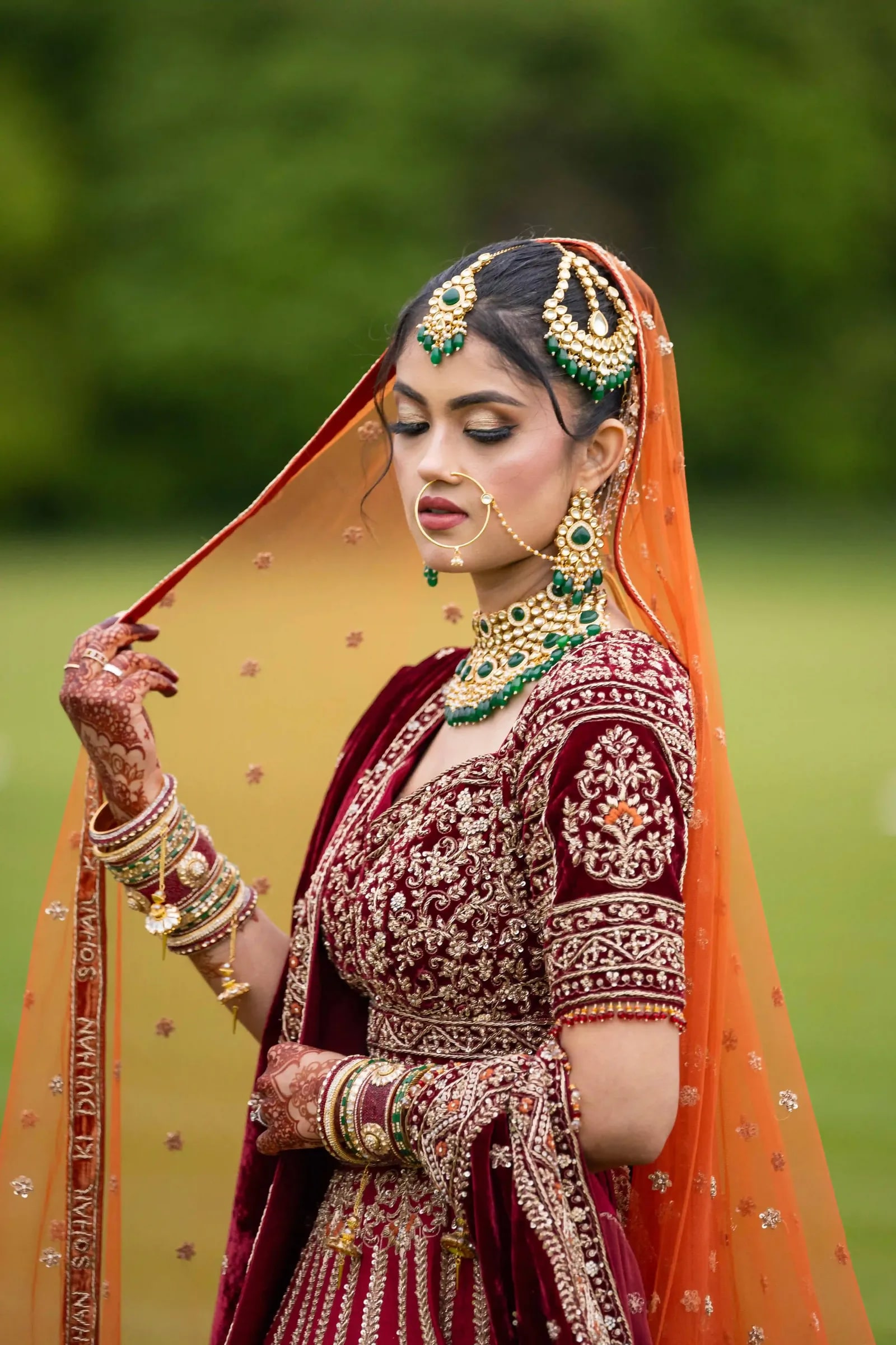 Bride holding her orange dupatta beside her face, in emerald and gold jewellery
