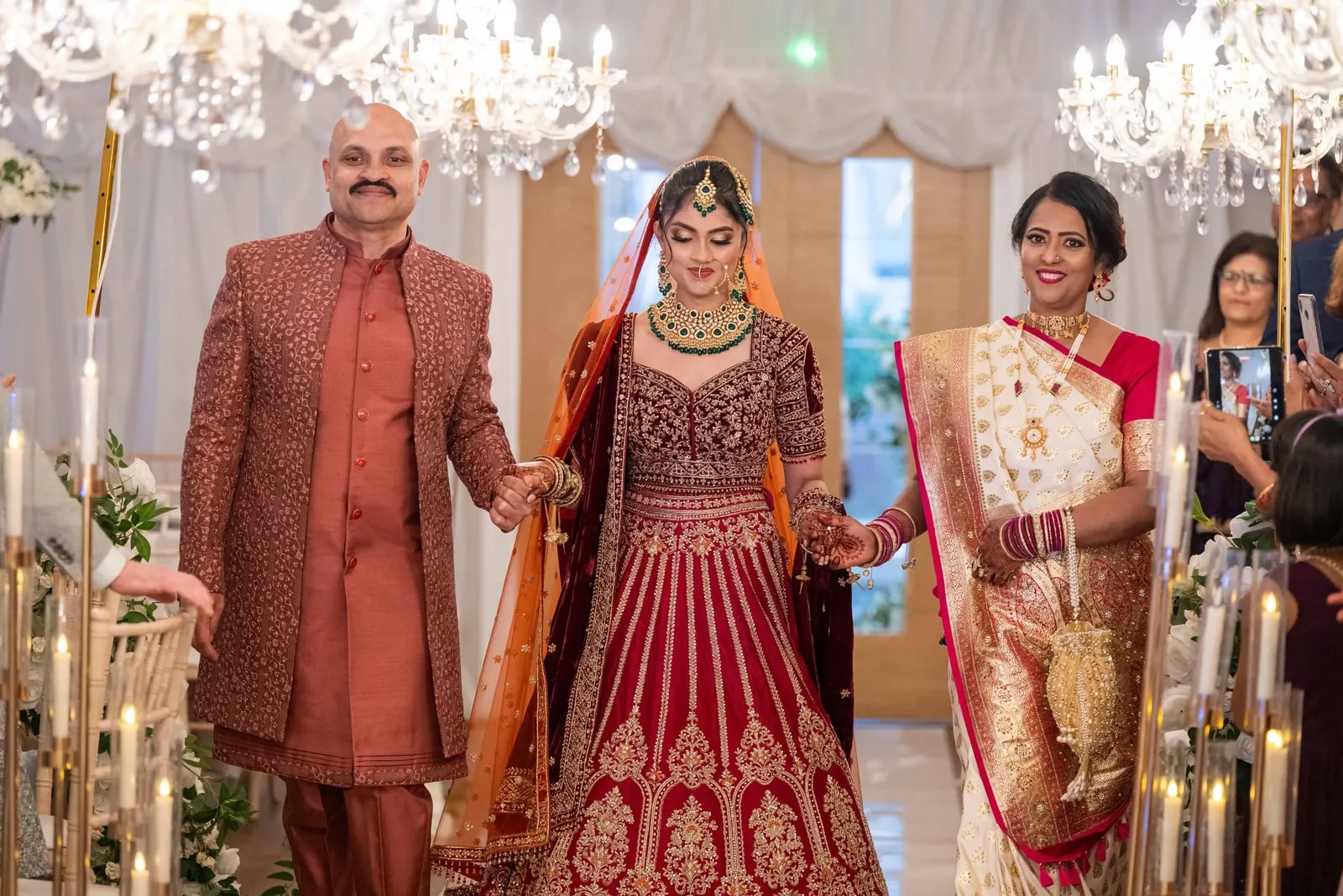 Bride walked in by her parents beneath chandeliers, both holding her hands