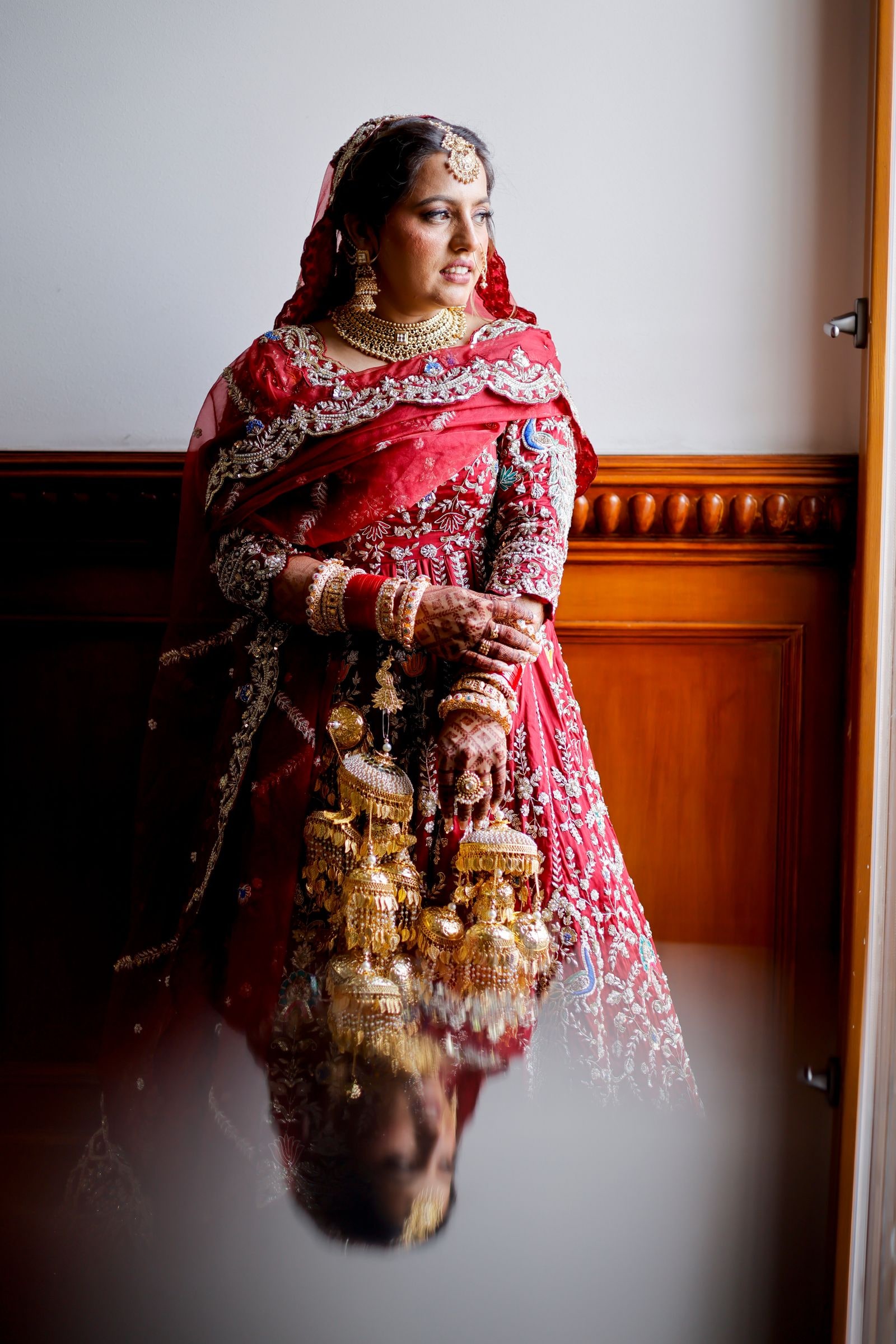 Bride in a red lehenga holding her gold kalire against a panelled wall