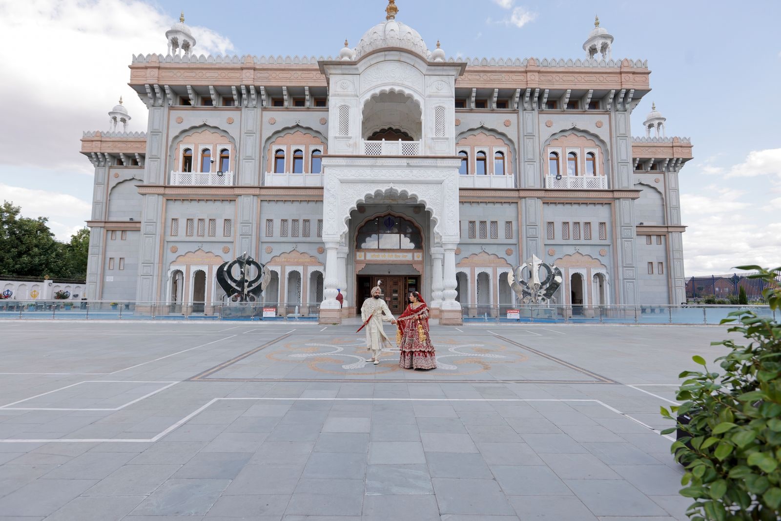 The full white front of the gurdwara, the couple tiny on the empty forecourt