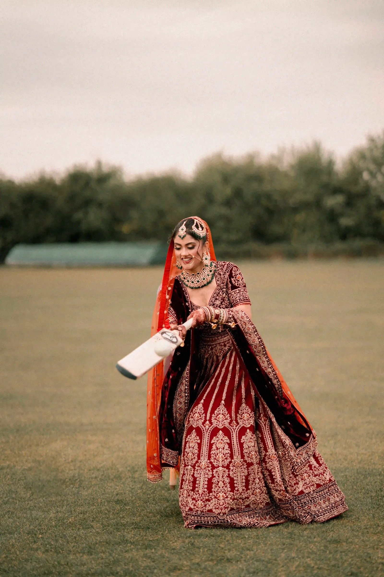 Bride walking out to bat on the cricket field, bat in hand, in a red lehenga