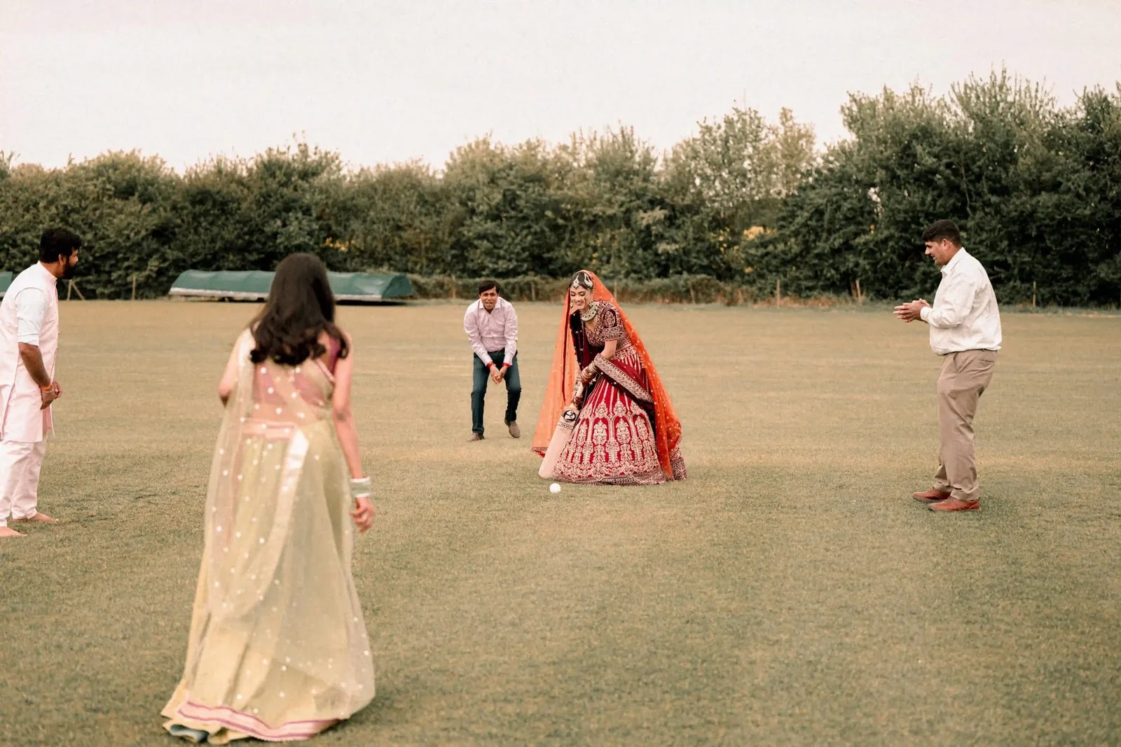 Guests fielding on the cricket outfield while the bride bats in her lehenga