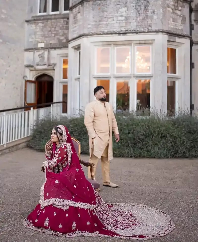 Bride seated on the terrace at Froyle Park in a crimson lehenga, her groom walking behind