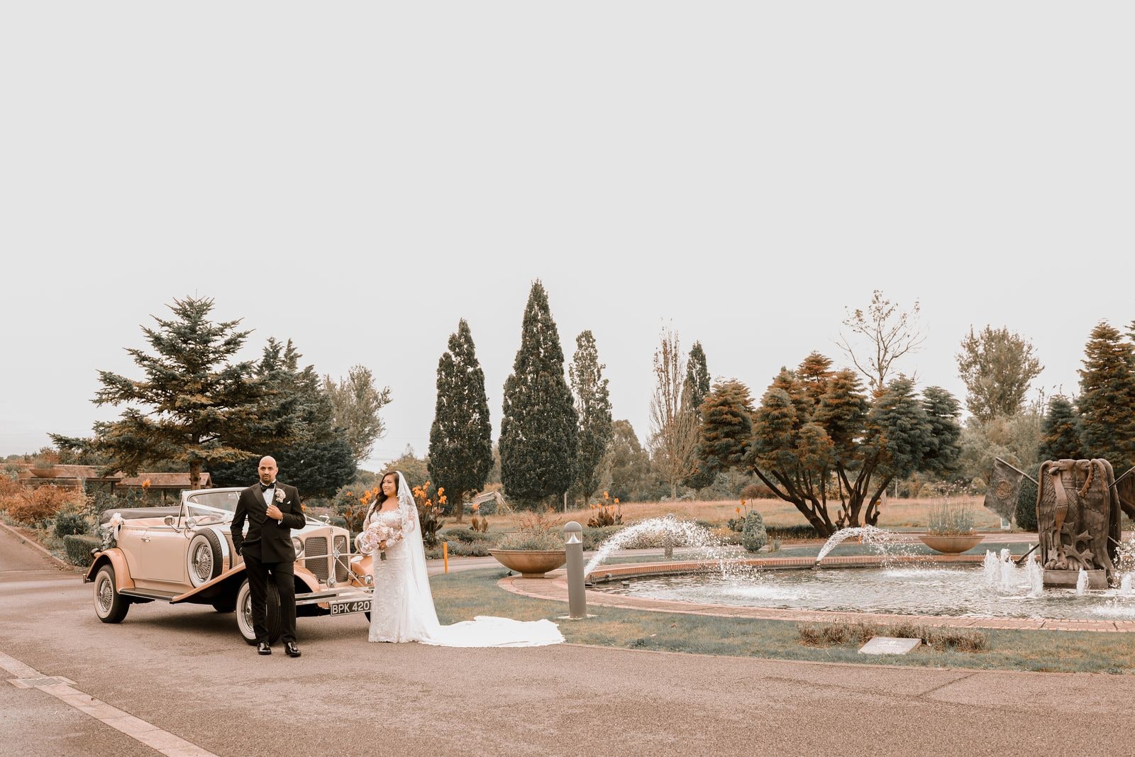 Bride and groom beside the vintage car by the fountains, her train across the tarmac