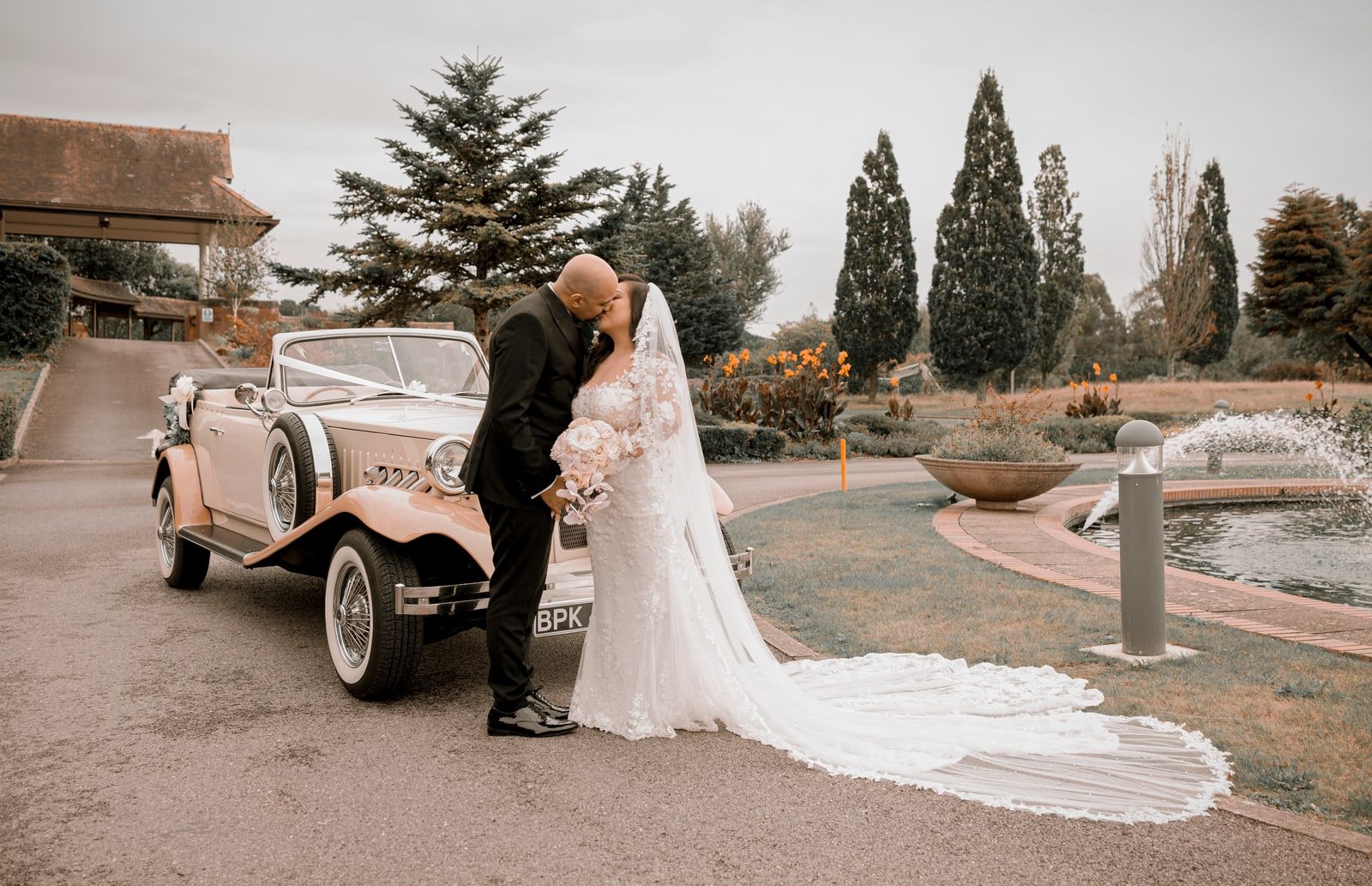 Groom kissing his bride beside the vintage car, the fountains playing behind