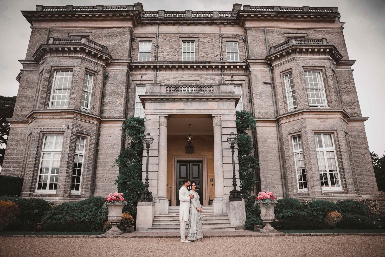 Couple standing small in the pillared doorway of Hedsor House, the grey stone front rising above them