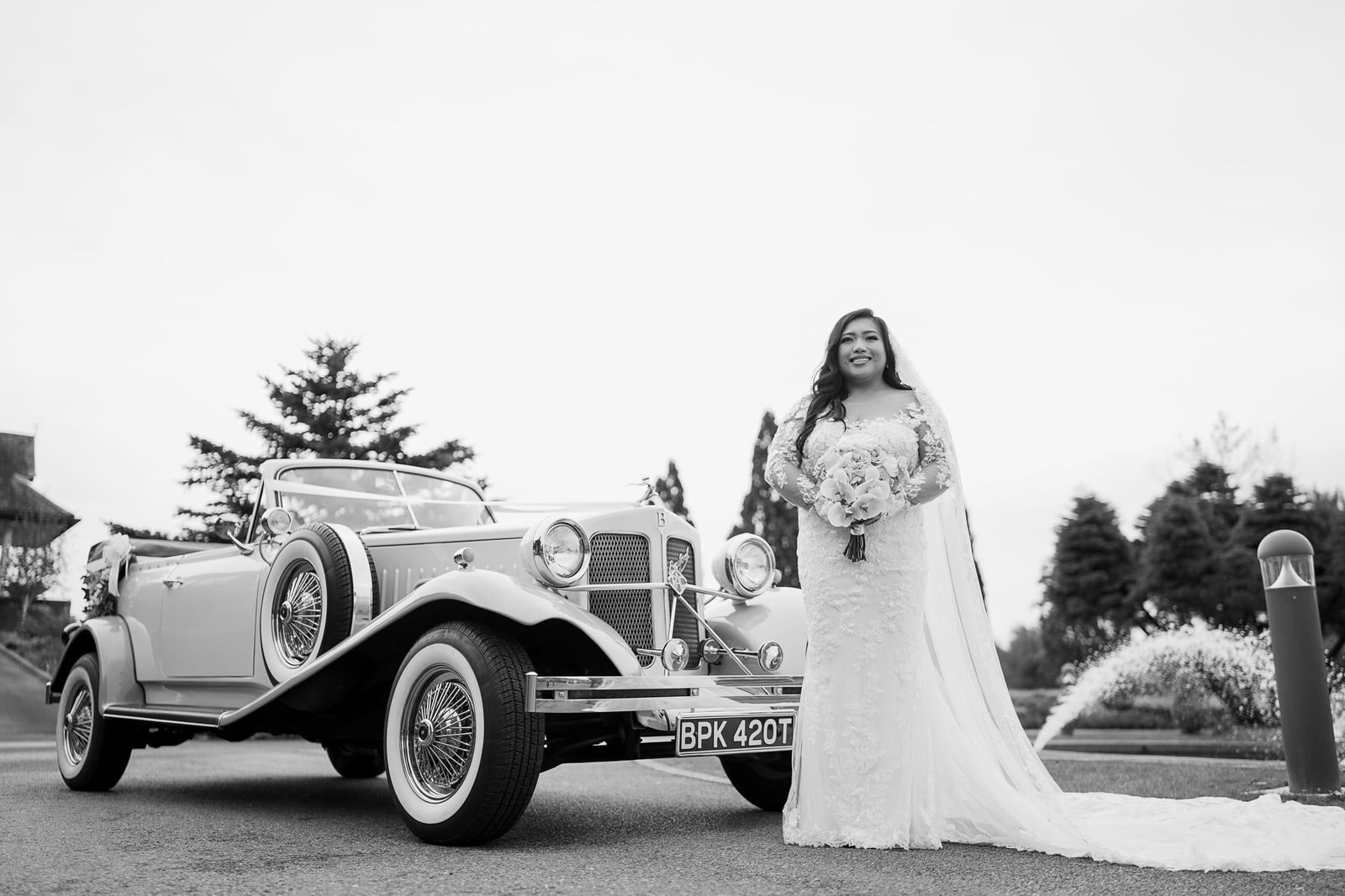 Bride standing alone beside the vintage Beauford, her train spread out