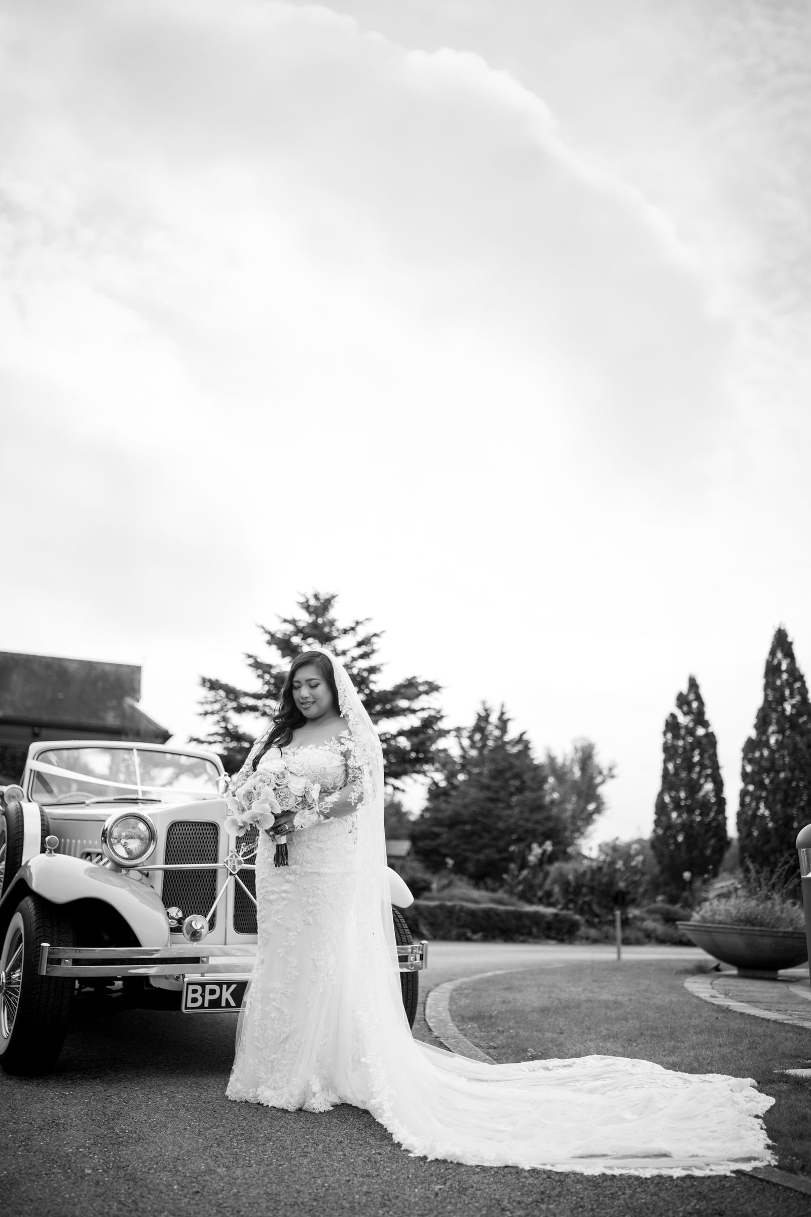 Bride beside the vintage car under a big cloudy sky, her train laid out behind, in black and white