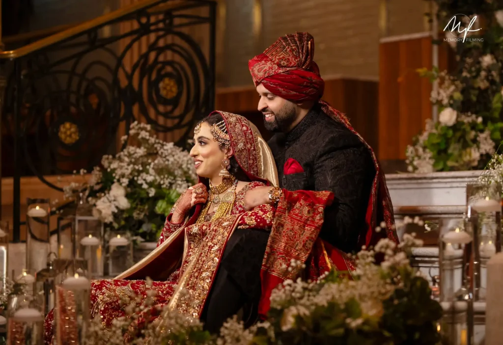 Bride in red and gold bridal lehenga with groom in red turban and black sherwani, indoor wedding ceremony with floral decor for asian wedding photography and videography packages London