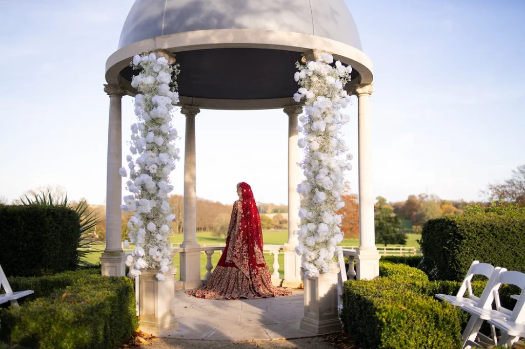 A Muslim wedding photography of a bride in london in a red and gold bridal lehenga standing under a rose-covered garden gazebo