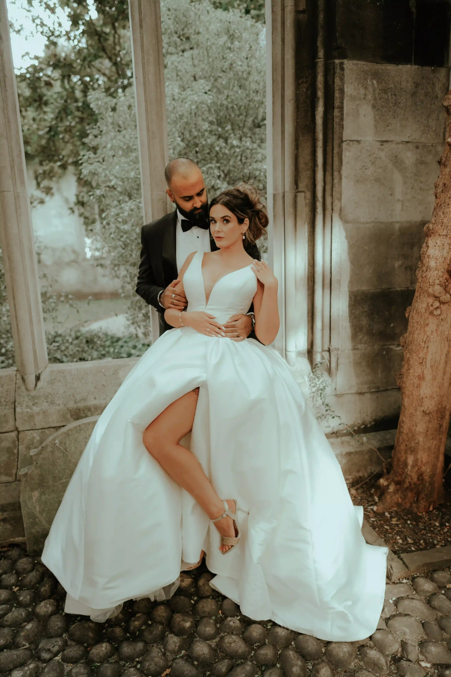 Bride in white ball gown with leg reveal embraced by groom under a stone archway, London wedding photography