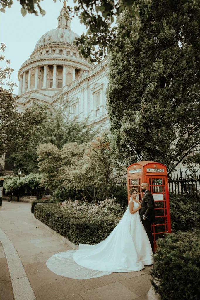 Bride and groom standing beside a classic red telephone box with a historic domed building in the background, London wedding photography