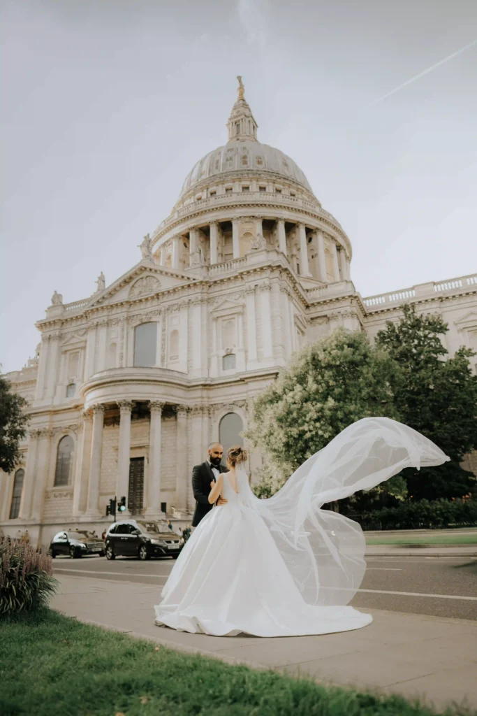 Bride's veil flowing in the wind as she stands with groom in front of a grand historic dome, London wedding photography